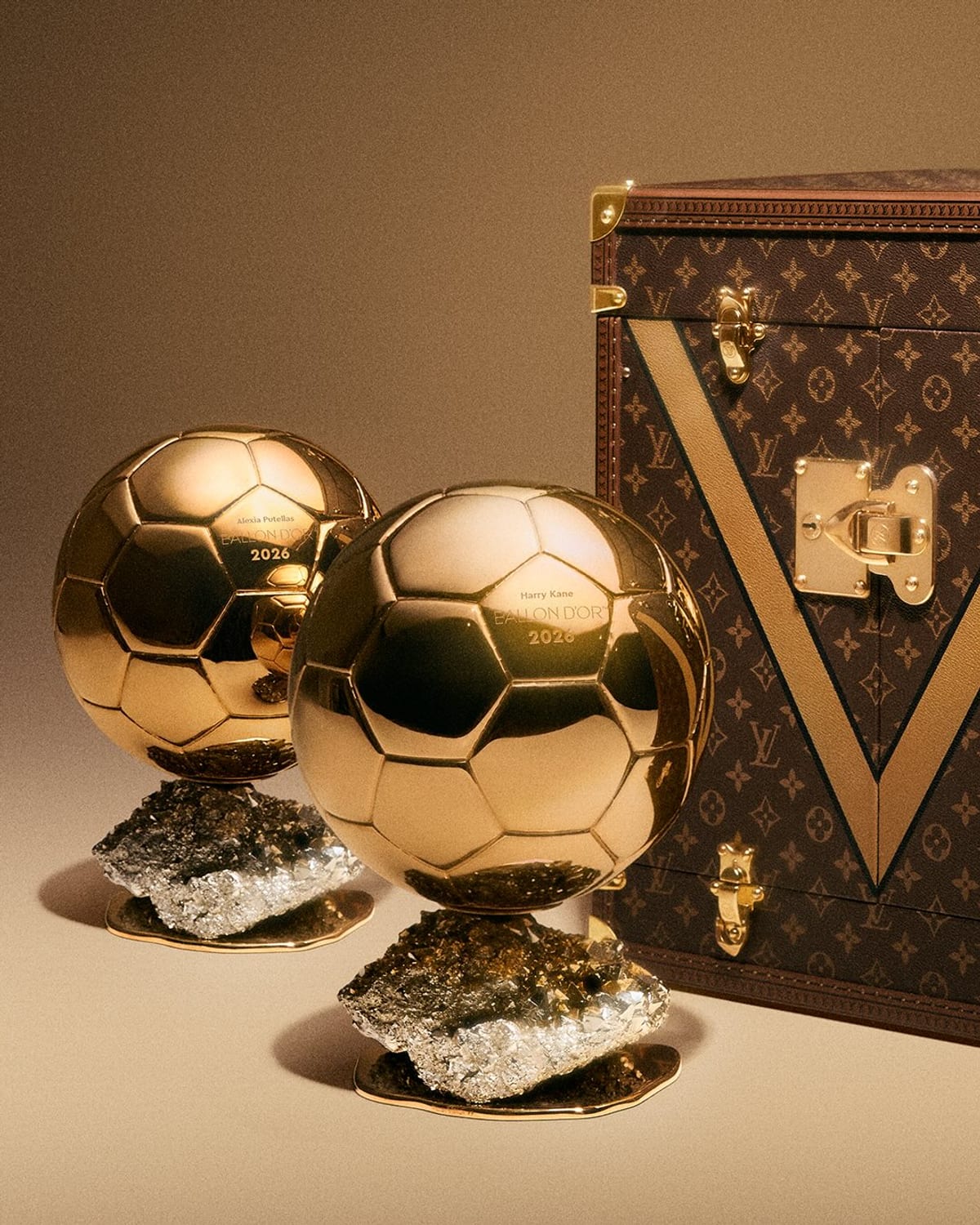 Two golden Ballon d'Or trophies, each a gold football on a base of rough pyrite, in front of a brown Louis Vuitton Monogram trunk with a large gold painted V across its front and a brass lock
