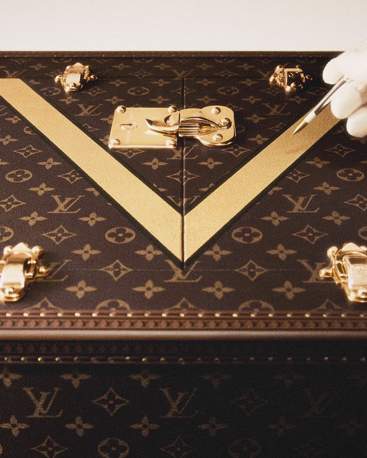 A close view of the lid of a Louis Vuitton Monogram trunk with a broad gold V painted across it and a gloved hand drawing a fine line along its edge with a pointed tool, brass corner clasps at each side and a brass lock at the centre