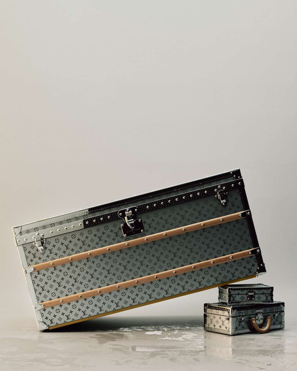 A large silver Louis Vuitton trunk engraved with the Monogram in black, with riveted steel edges, two tan leather slats and a steel lock, tilted on a wet floor beside two small matching cases stacked on top of each other