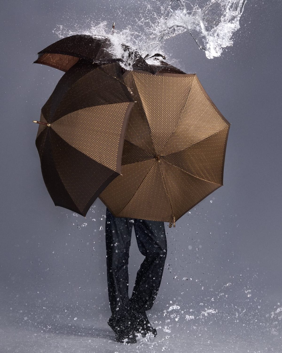 Three brown Louis Vuitton Monogram umbrellas held up against a burst of splashing water, hiding the person carrying them down to the legs, in dark trousers and dark shoes on a wet grey floor