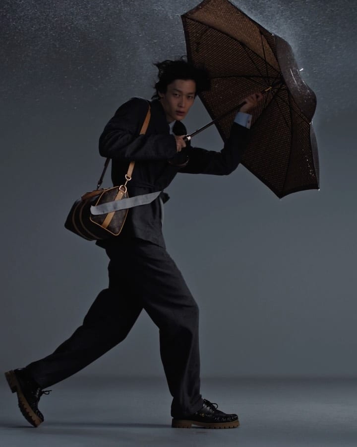 A model in a dark grey suit and black lug-sole shoes leaning into the wind under a brown Monogram umbrella, a brown Monogram bag slung across his body, against a grey backdrop with spray