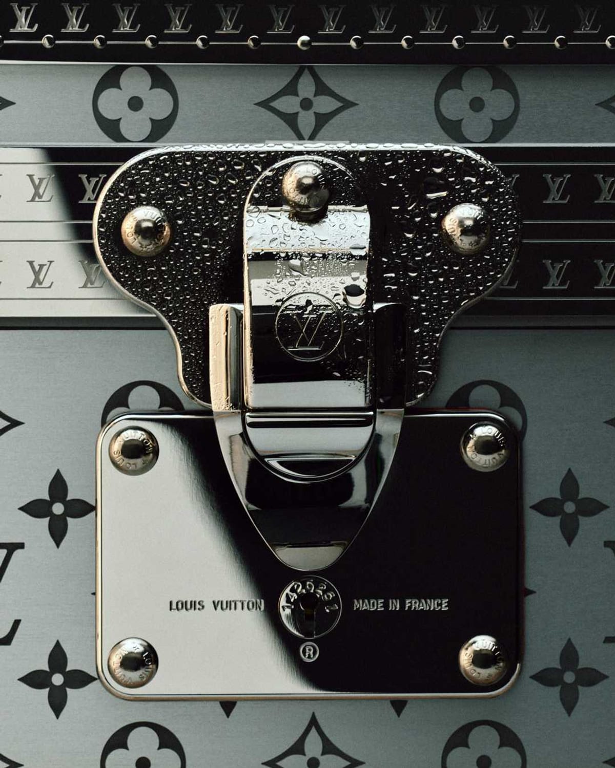 Close-up of a Louis Vuitton trunk lock beaded with water drops, a polished steel clasp and plate stamped Louis Vuitton and Made in France against the black-on-silver Monogram and a row of LV-stamped trim