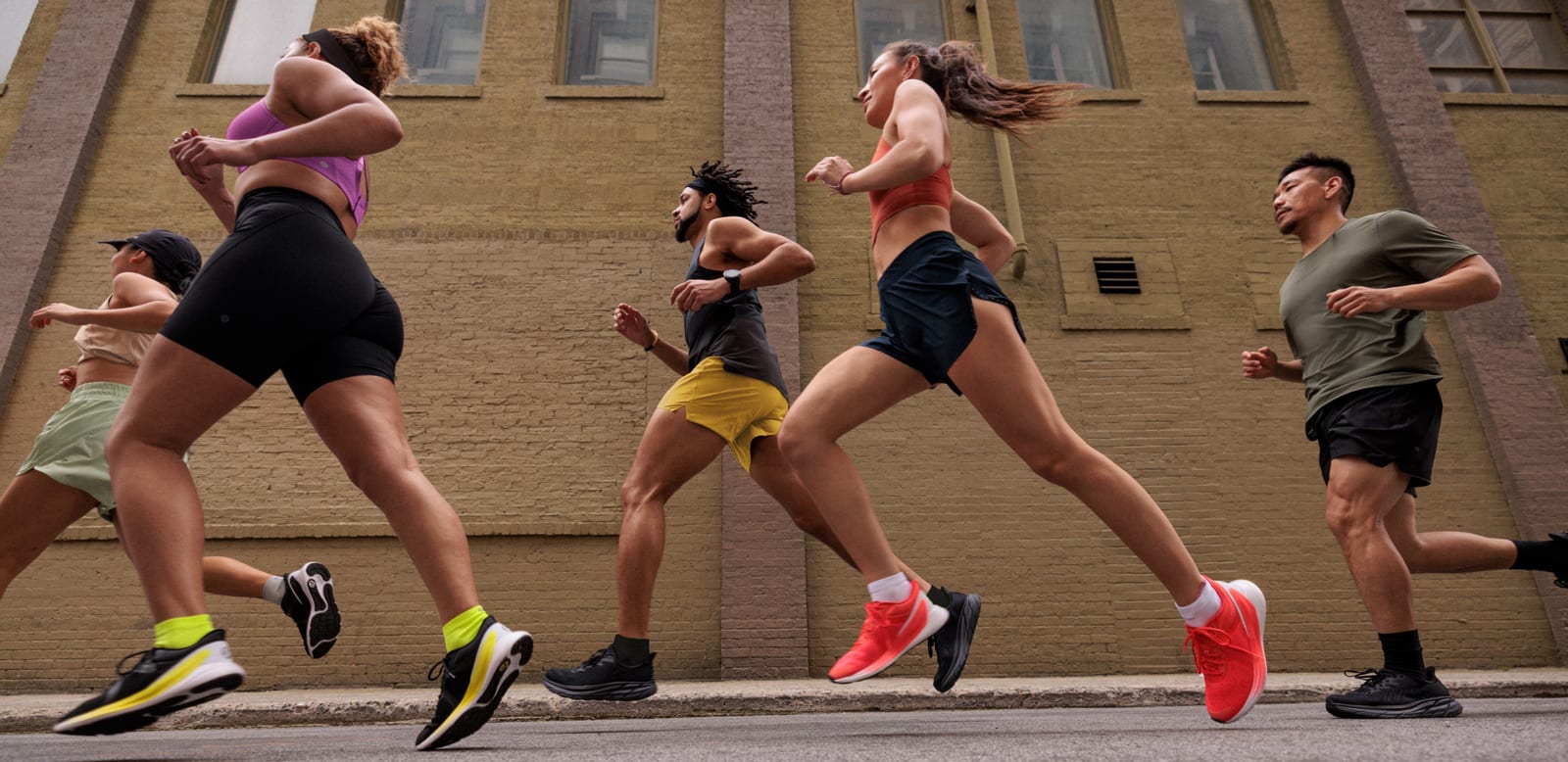 Five runners in lululemon shorts, tanks and tees mid-stride on a city street past a brick building, wearing lululemon running shoes in black, coral and yellow