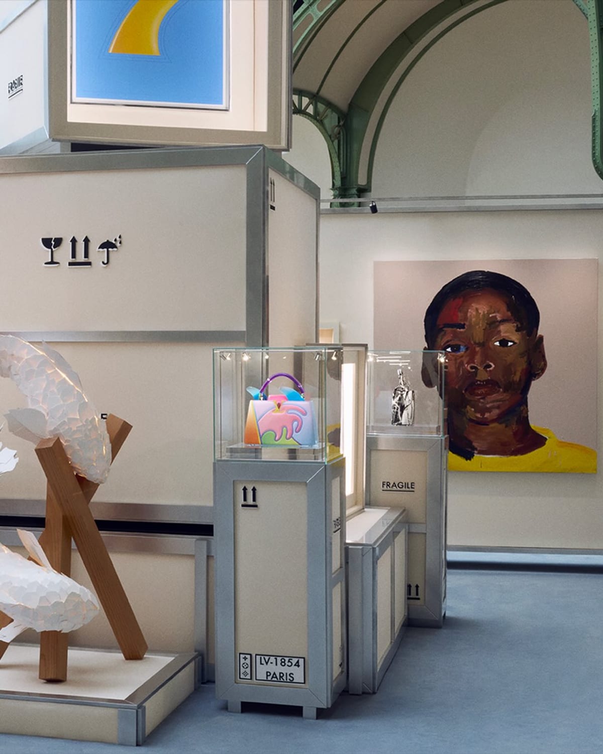 A rainbow-coloured Capucines bag in a glass vitrine on a trunk-like plinth stencilled LV-1854 Paris, beside a white sculpture on wooden supports, with a large painted portrait of a man in a yellow top on the wall behind, under the Grand Palais's green ironwork