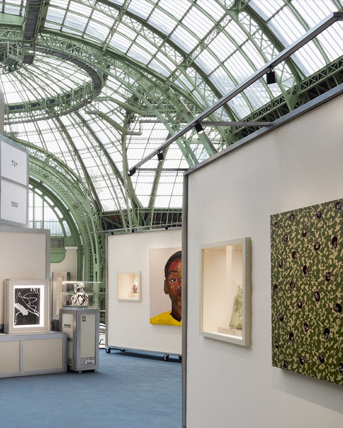Louis Vuitton's Artycapucines retrospective under the glass roof and green ironwork of the Grand Palais: white display walls hung with a portrait of a man in a yellow top and a green panel with dark spots, bags in vitrines on trunk-like plinths