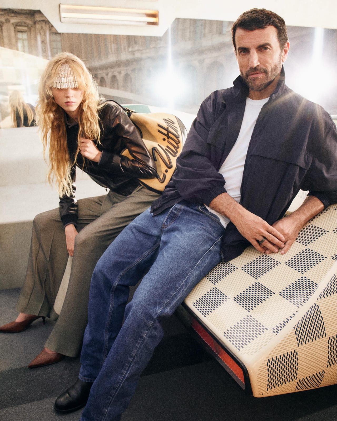 Nicolas Ghesquière in a navy jacket and jeans sitting with model Ella Dalton on a Damier-patterned concept car in the Cour Carrée du Louvre, Louis Vuitton's portrait from the Spring 2027 show