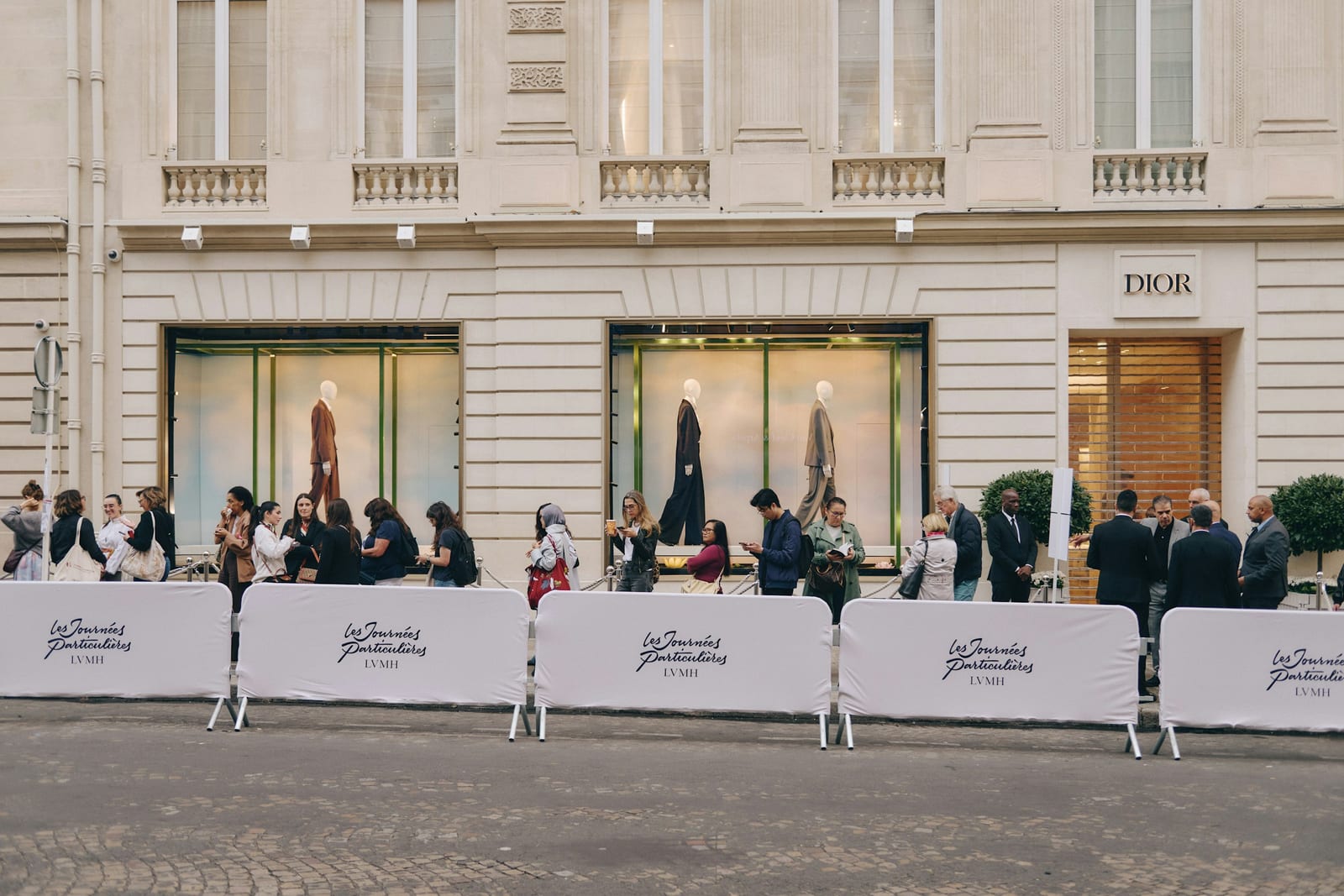 A line of visitors waits behind white barriers printed with Les Journées Particulières LVMH outside a cream stone building with a Dior sign, two shop windows showing mannequins in tailoring against a pale sky