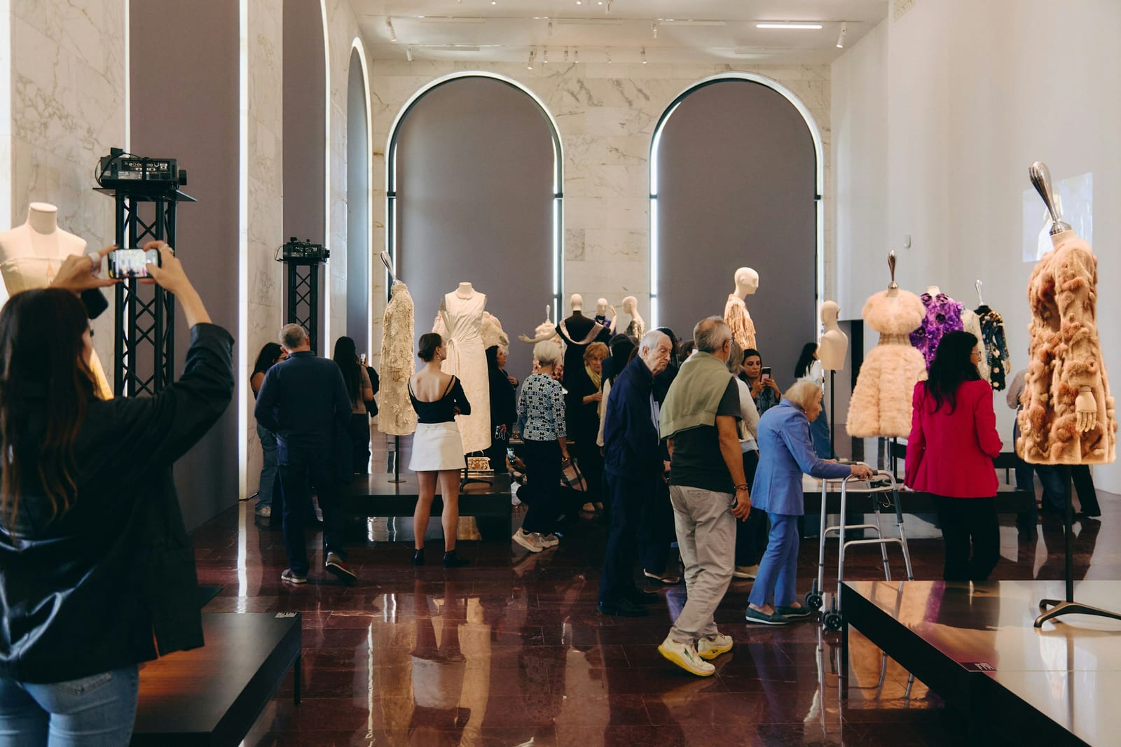 Visitors move through a marble hall with two tall arched windows, looking at dresses and fur coats on mannequins, one a pale pink ruffled dress and one a tufted tan fur coat