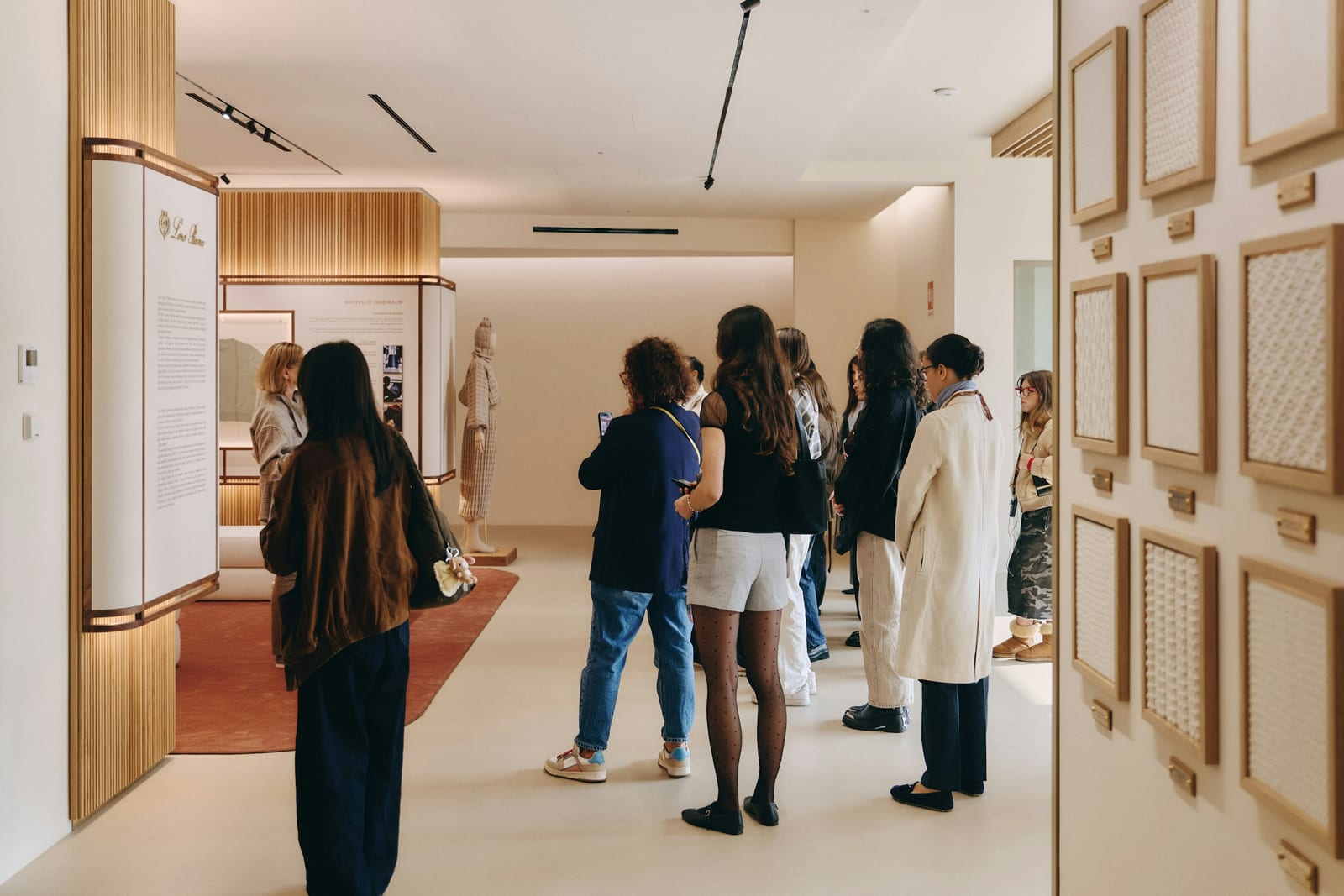 Visitors stand in a pale, wood-panelled exhibition room with a Loro Piana text panel on the left, a knitted garment on a mannequin at the back and framed swatches of knitted fabric on the right wall