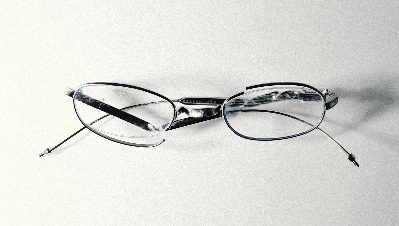 A pair of thin silver metal frames from the Maison Margiela and Gentle Monster collection on a pale grey background, the left rim split open and both lenses scratched, with a row of numbers engraved across the bridge