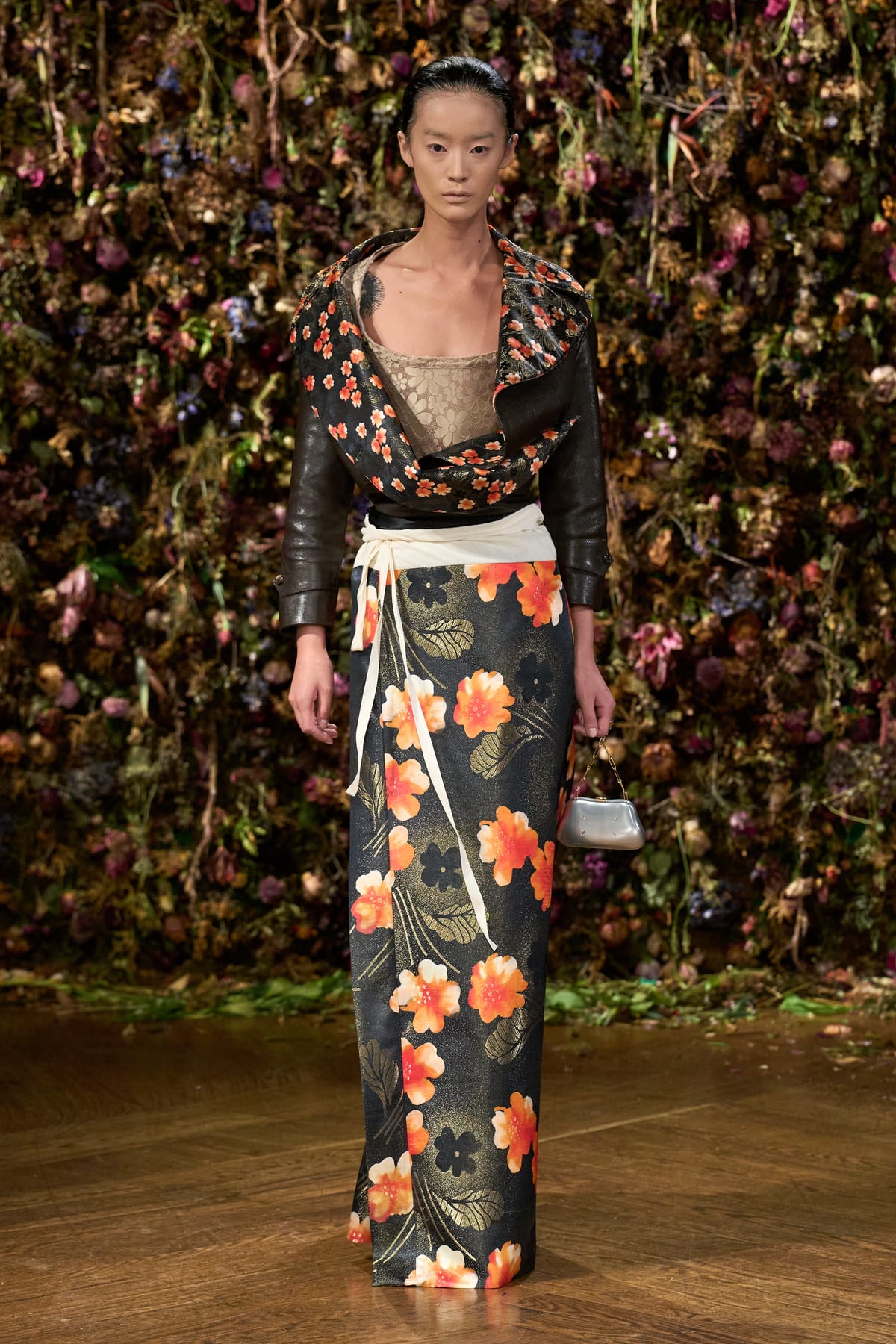 A Maison Margiela Spring/Summer 2027 look: a black leather jacket with a floral-print collar over a long skirt printed with orange flowers, with a cream sash at the waist and a small silver bag