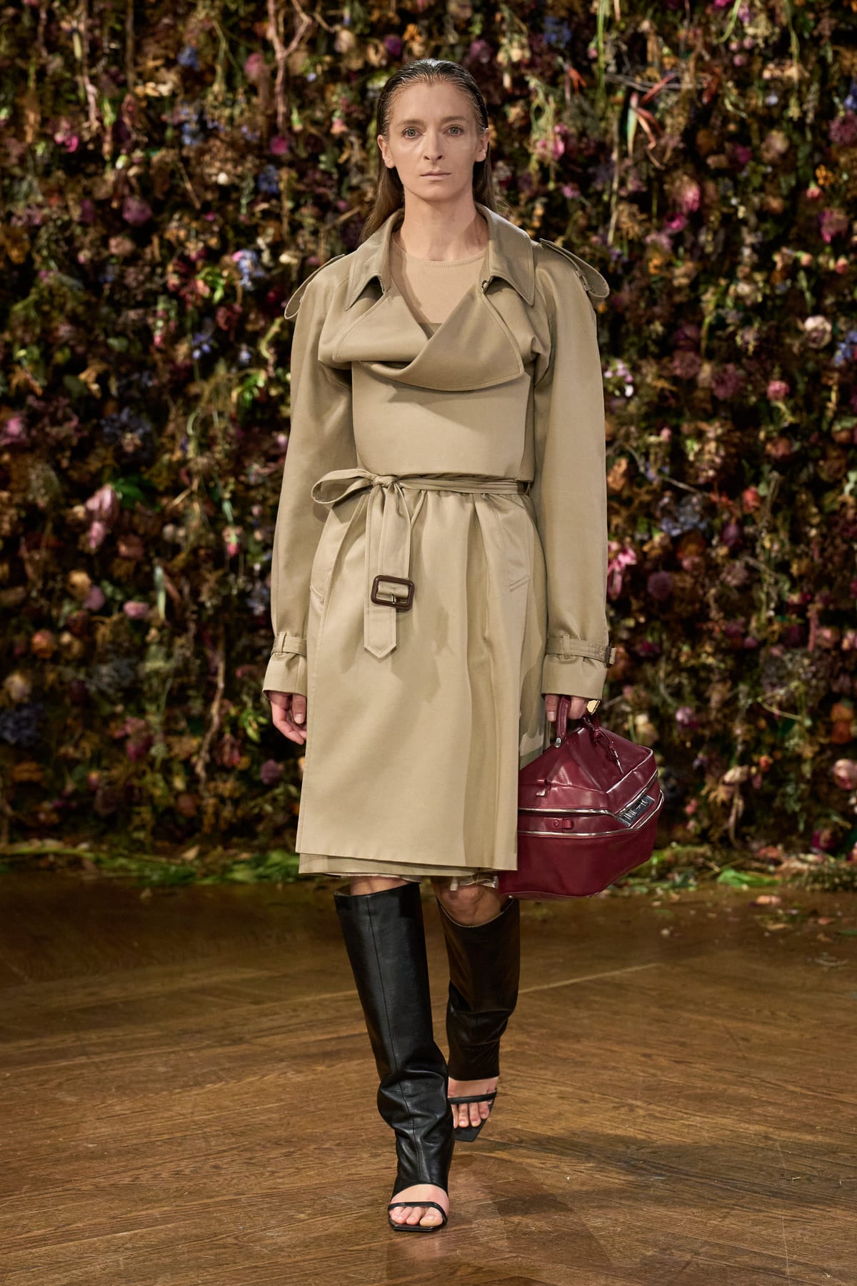 Look 1 from Maison Margiela Spring/Summer 2027: a khaki trench coat with a draped, cowl-like front, belted, worn with black knee-high boots and a burgundy bag in front of a wall of dried flowers
