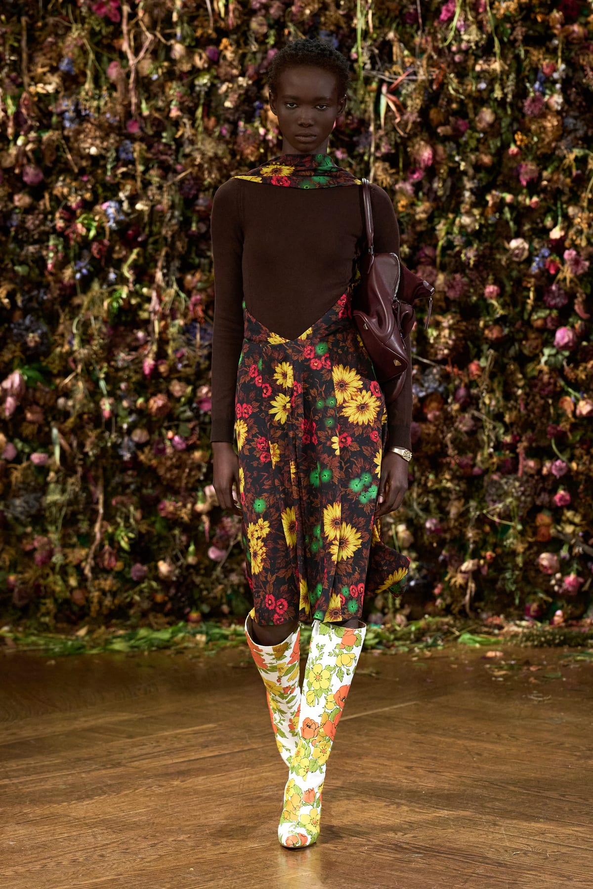 A Maison Margiela Spring/Summer 2027 look: a brown knit top and scarf with a sunflower-print skirt and floral-print knee-high boots, with a burgundy bag