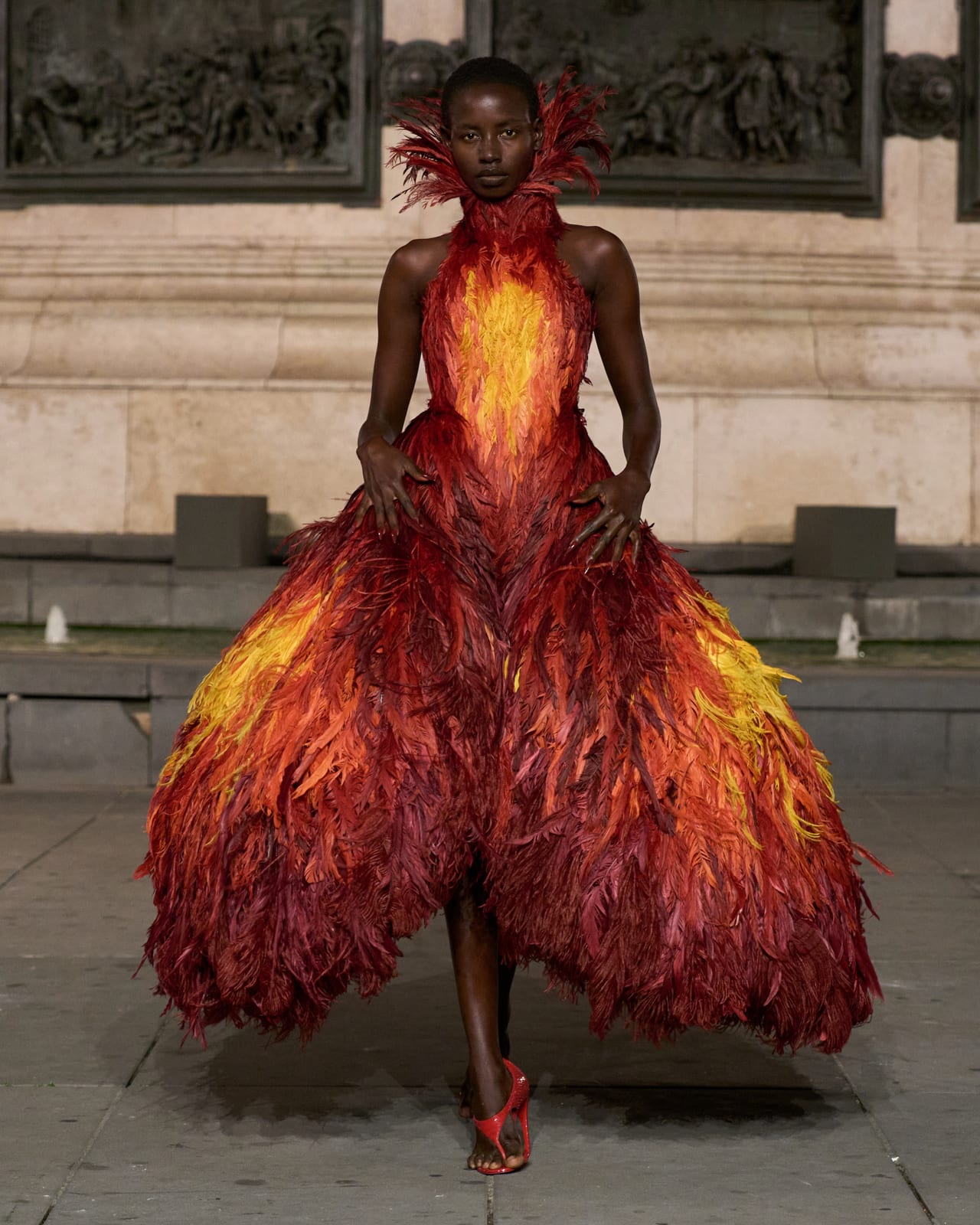 A Matières Fécales Spring/Summer 2027 look at Place de la République: a feathered gown shading from red to yellow like a flame, with a feathered collar