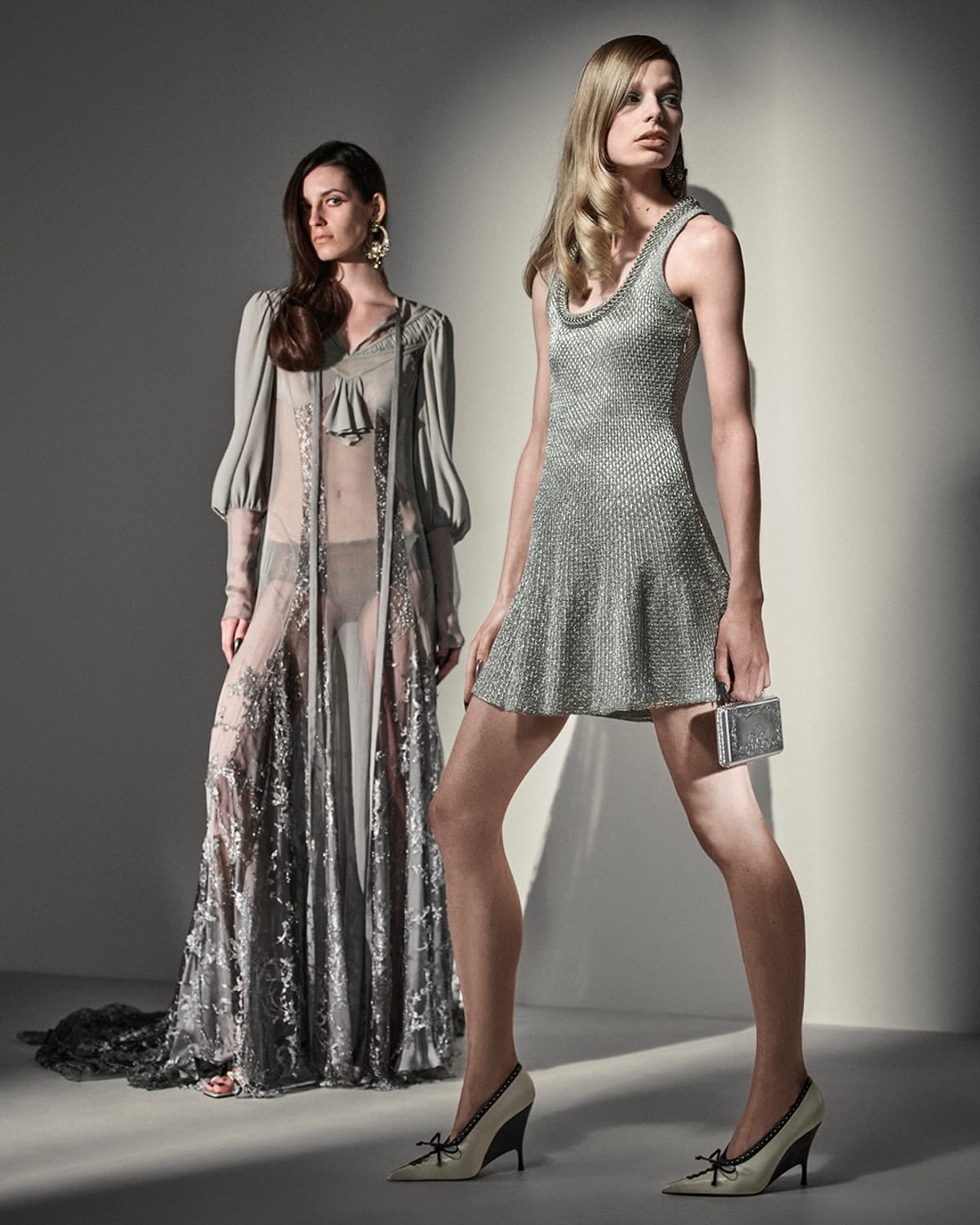 Two models against a grey wall: one in a long sheer grey gown with billowing sleeves and silver embroidery, the other in a short sleeveless silver metallic knit dress, cream bow-trimmed pumps and a small silver box clutch