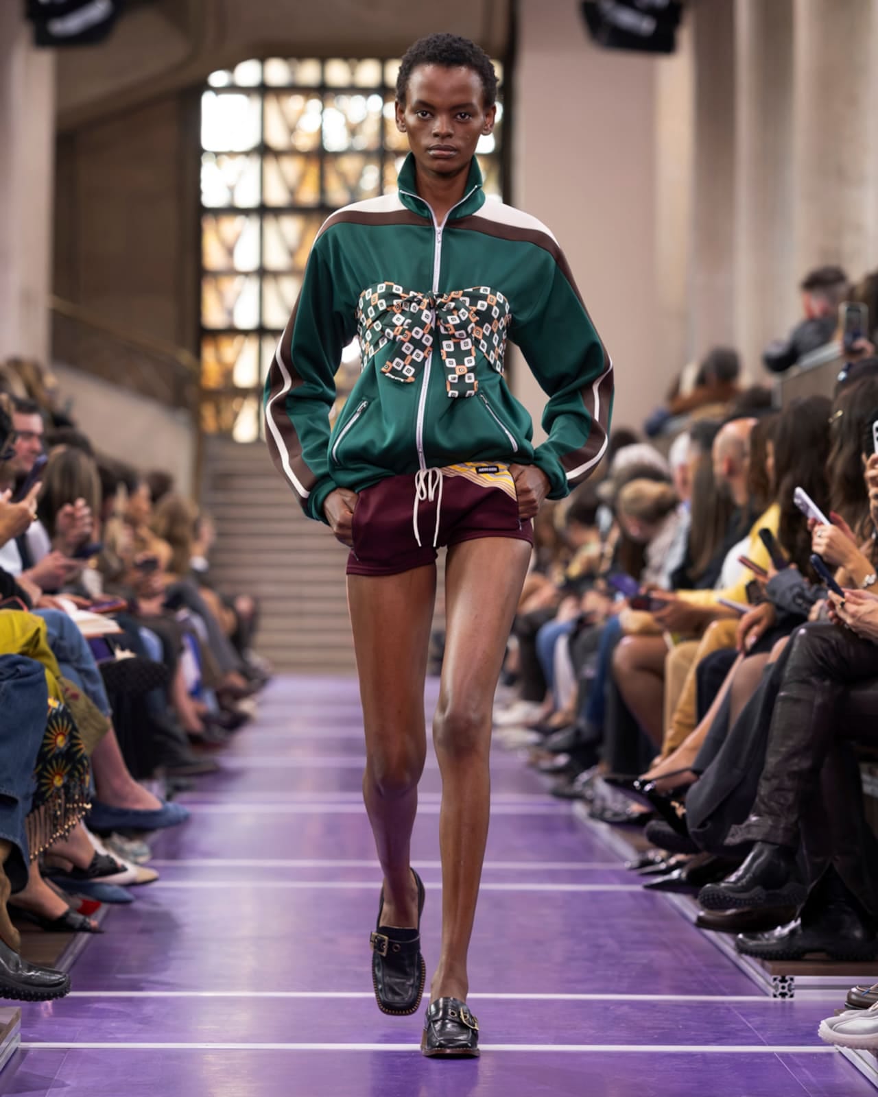 A Miu Miu Spring 2027 look: a green zip track jacket with a geometric-print bow-tied top over burgundy shorts and black buckled loafers