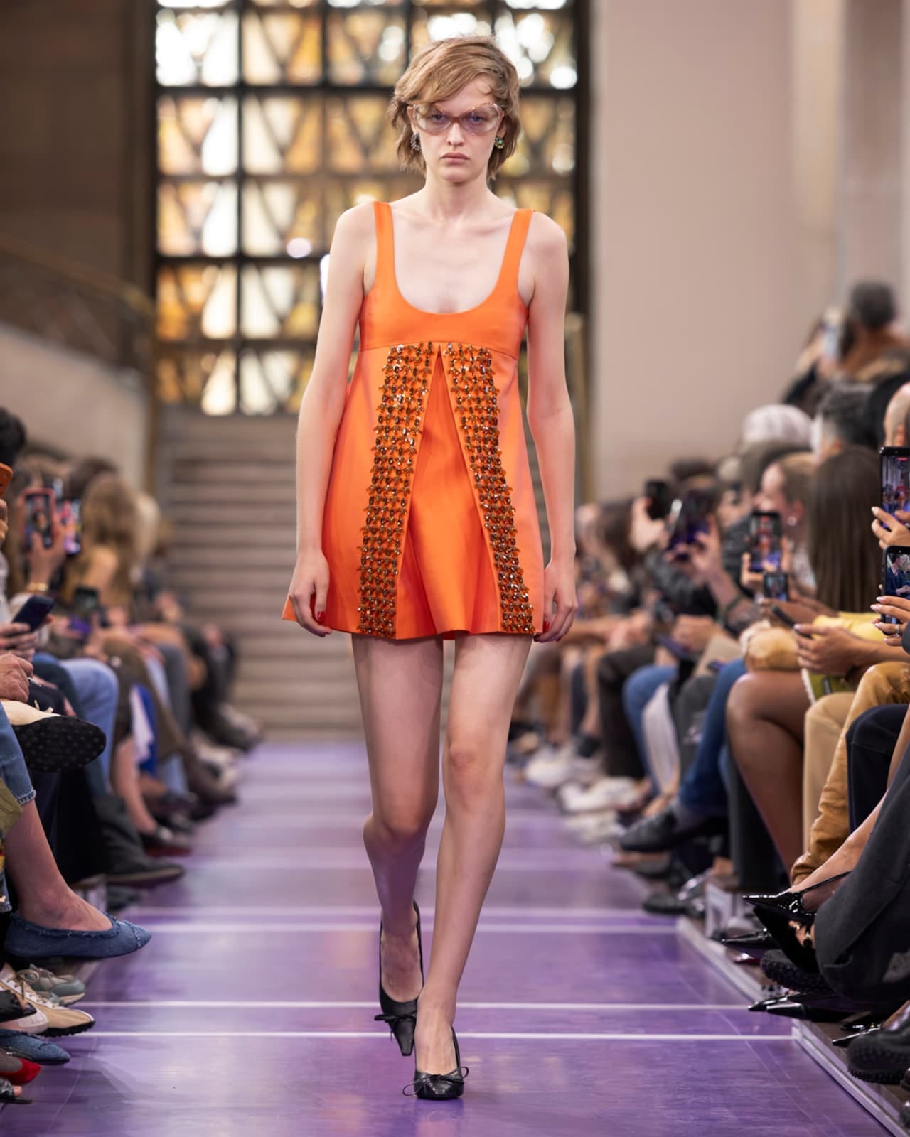 A Miu Miu Spring 2027 look: a bright orange sleeveless mini dress with a beaded front panel, worn with pale tinted glasses and black bow pumps