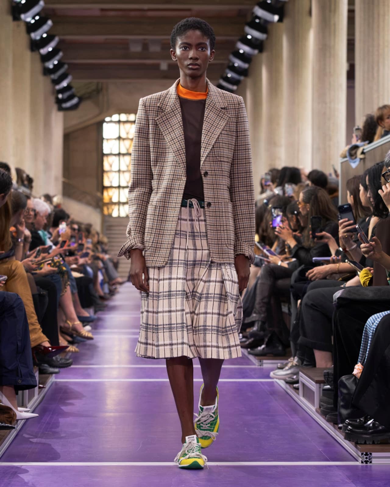 A Miu Miu Spring 2027 look: a brown checked blazer over a brown top with an orange collar and a checked drawstring skirt, worn with colourful sneakers on a purple runway