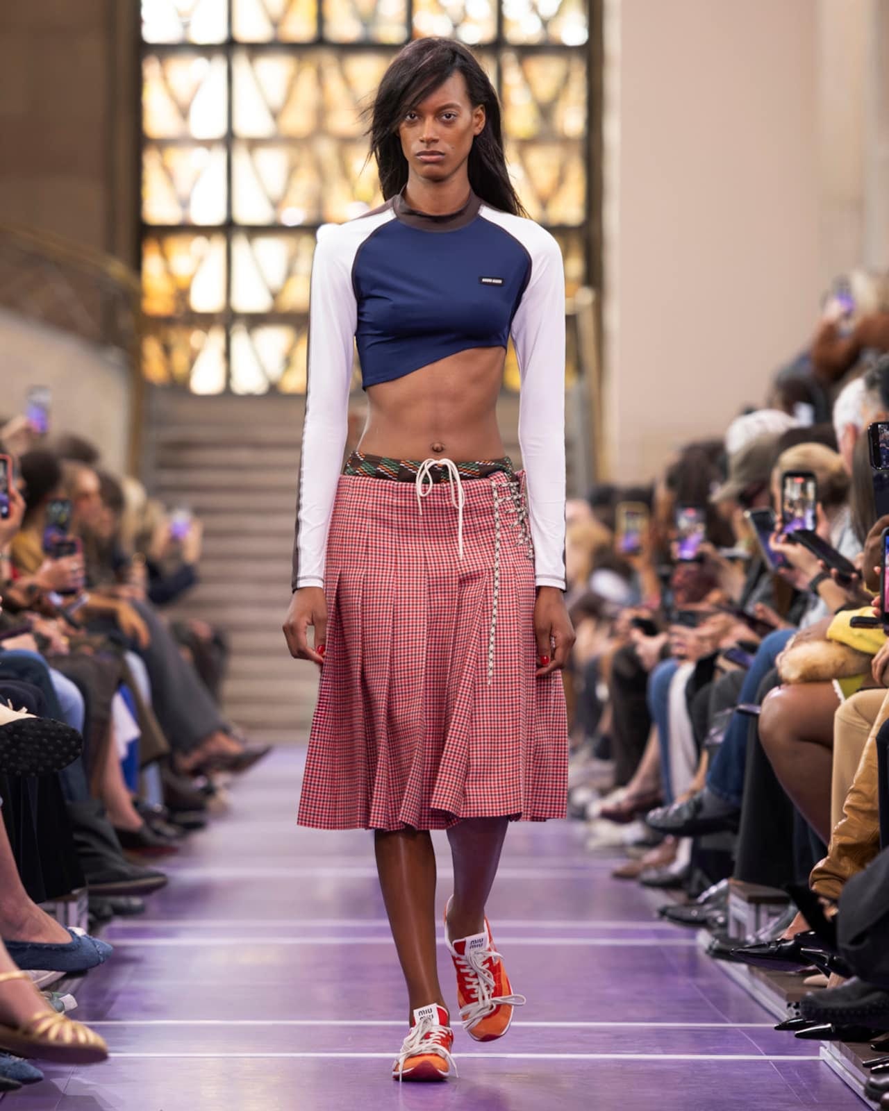 A Miu Miu Spring 2027 look: a navy cropped top with white raglan sleeves over a red checked pleated skirt, with a drawstring at the waist and bright sneakers