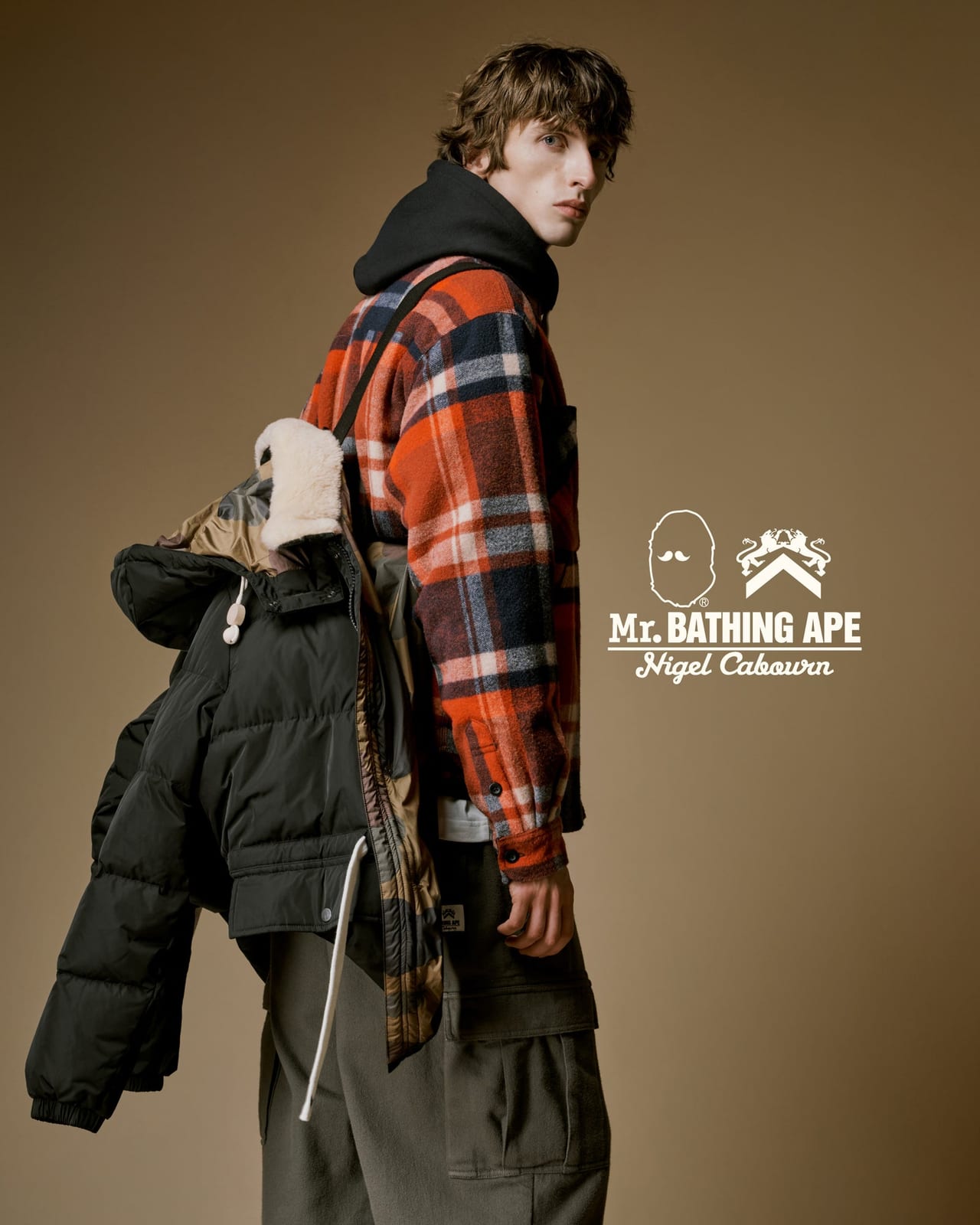 A model in an orange, navy and white check shirt jacket over a black hoodie, with a black down jacket lined in camouflage and trimmed with a cream shearling collar slung over one shoulder, beside the MR. BATHING APE and Nigel Cabourn logos