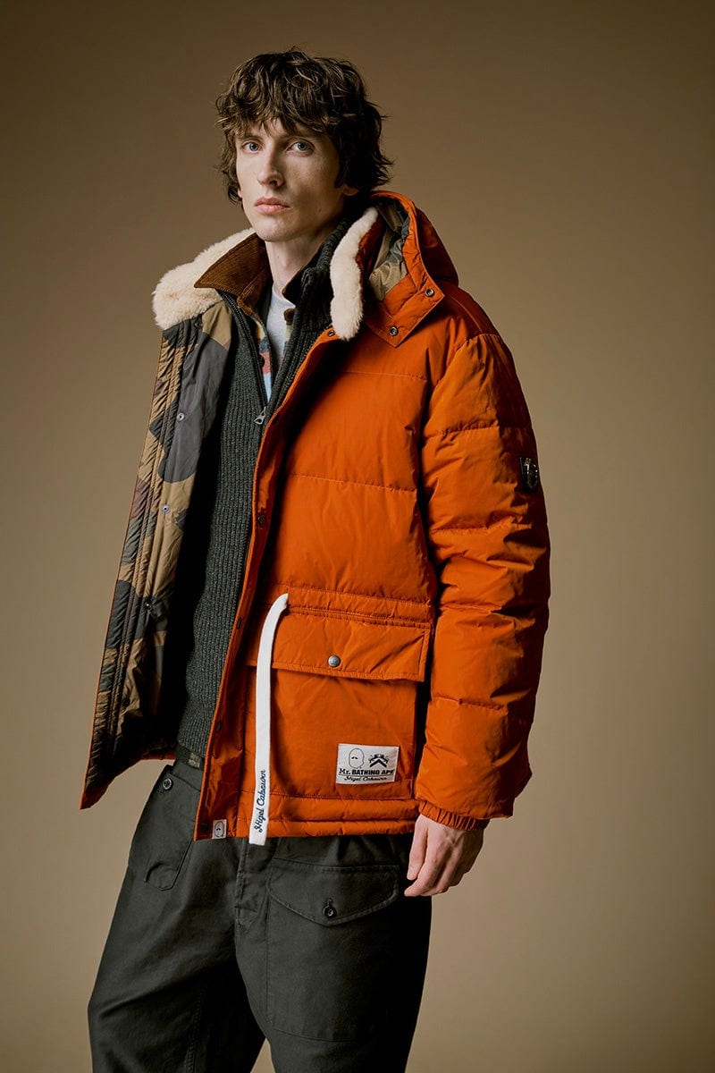 A model in a rust-orange hooded down jacket with a cream shearling collar, open to show its camouflage lining, a white tape printed Nigel Cabourn hanging from the pocket and a MR. BATHING APE and Nigel Cabourn label on it, over a charcoal ribbed zip-neck knit and olive trousers
