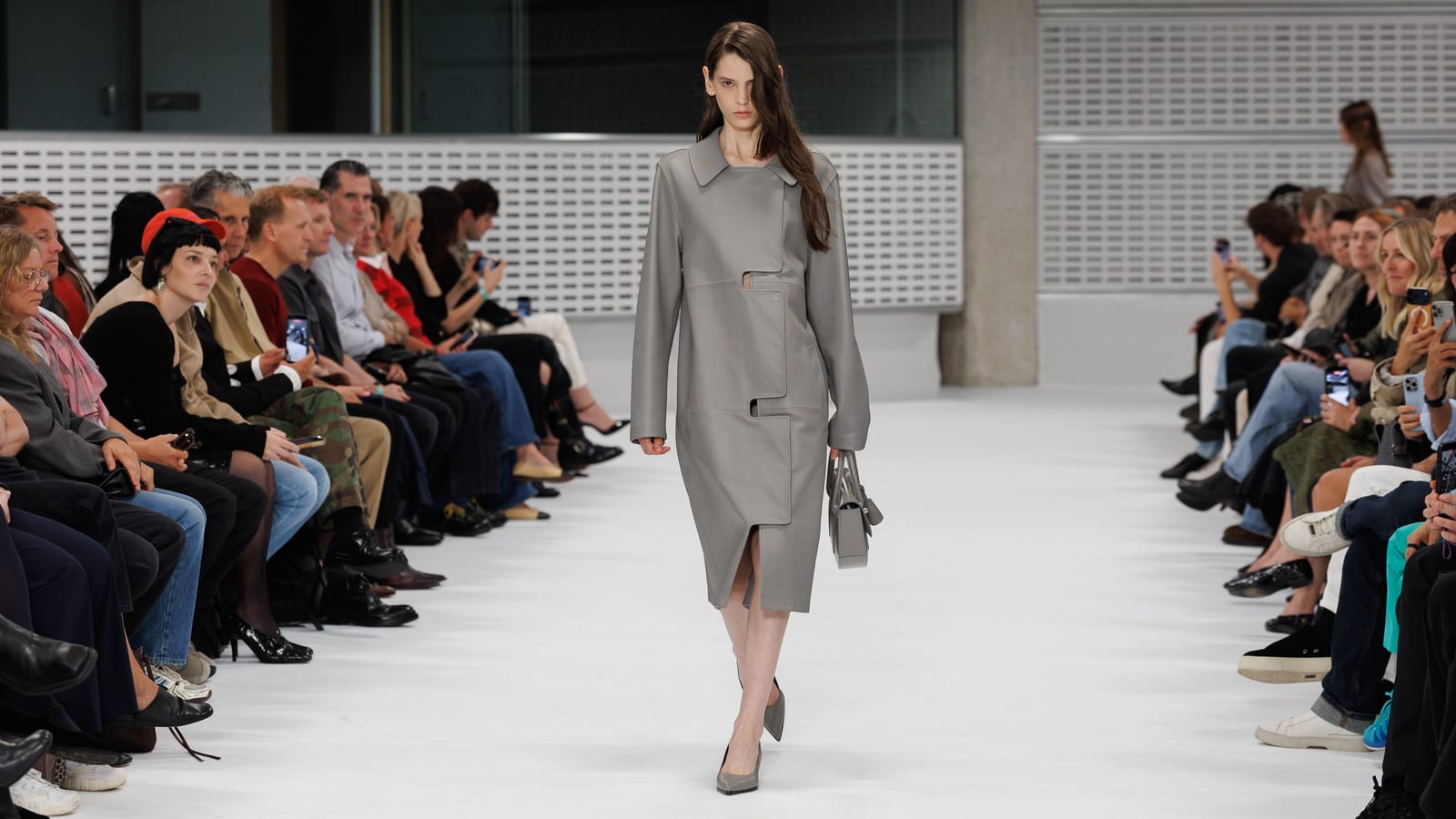 A model on the Mulberry by Christopher Kane Spring/Summer 2027 runway in a grey leather trench coat-dress with cutout paneling, carrying a matching grey leather bag