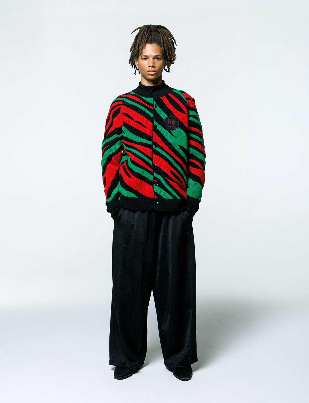A model in the Needles x A Tribe Called Quest Roll Neck Cardigan, knitted in jagged red, green and black stripes, with black Needles track pants