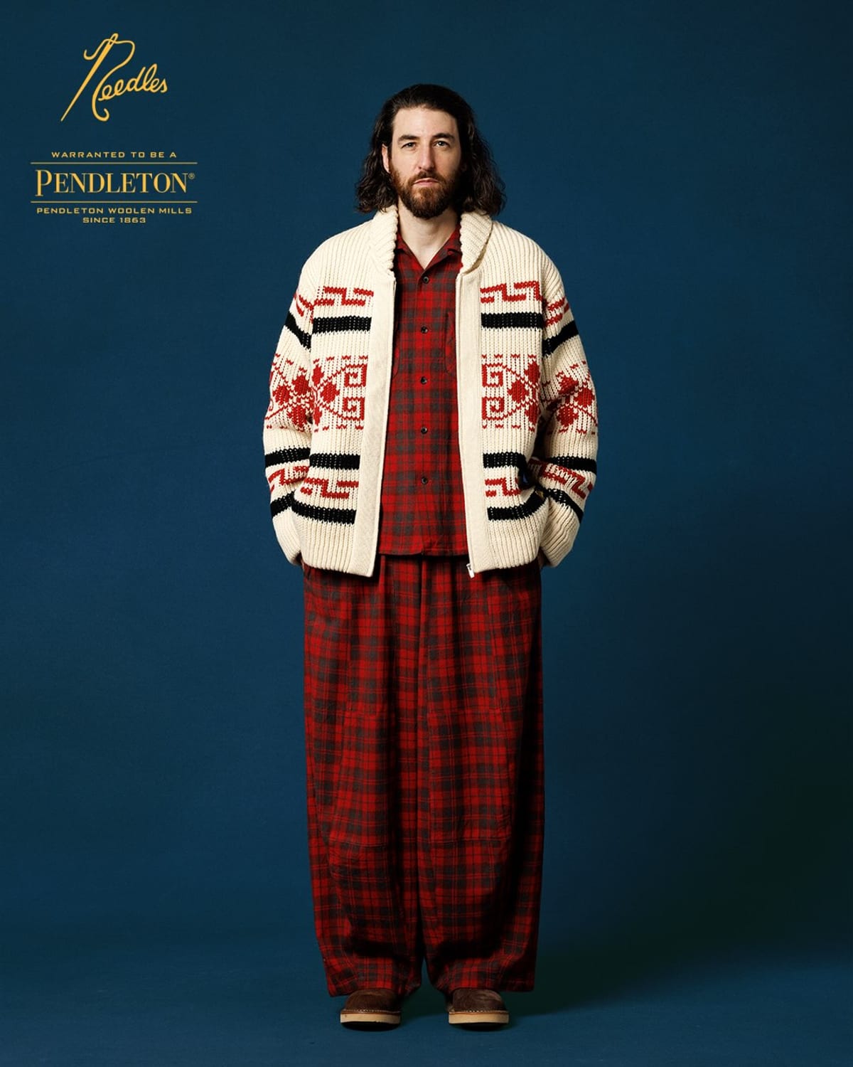 A model in the Needles x Pendleton Cowichan Sweater, a cream zip-up knit with a shawl collar and bands of red and black geometric pattern, over a red plaid shirt and trousers