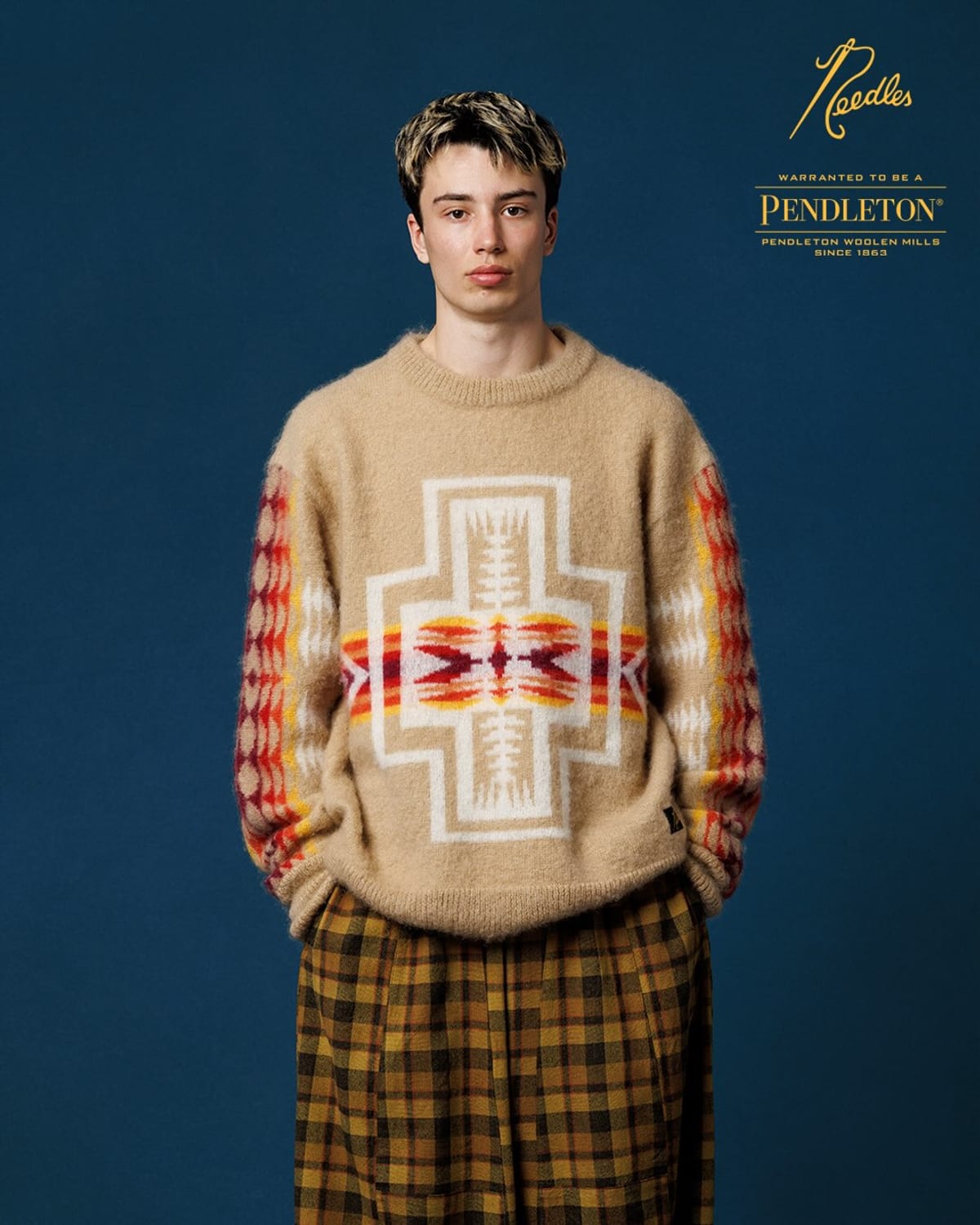 A model in the Needles x Pendleton Mohair Sweater, a fuzzy beige knit with Pendleton's Harding pattern in red, orange and cream across the chest and on both sleeves, worn with mustard plaid trousers