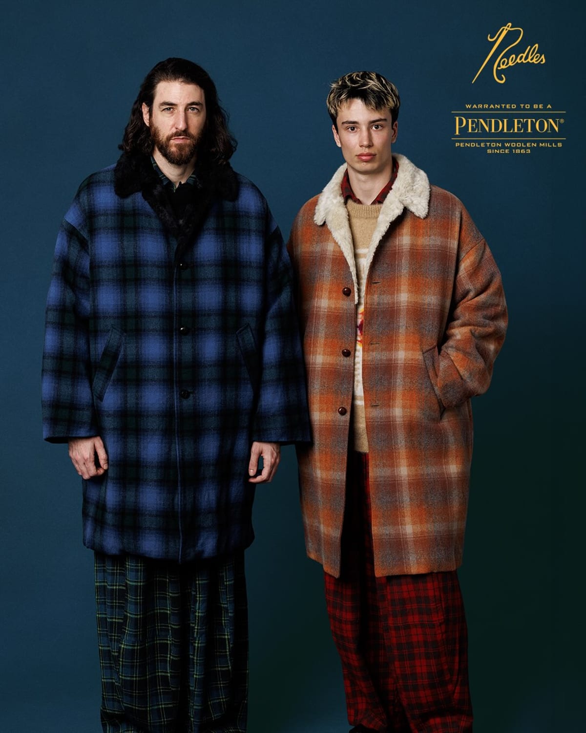 Two models against a navy backdrop in the Needles x Pendleton Shawl Collar Coat, one in blue, black and green plaid with a black fur collar, one in orange and grey plaid with a cream fleece collar, over plaid trousers