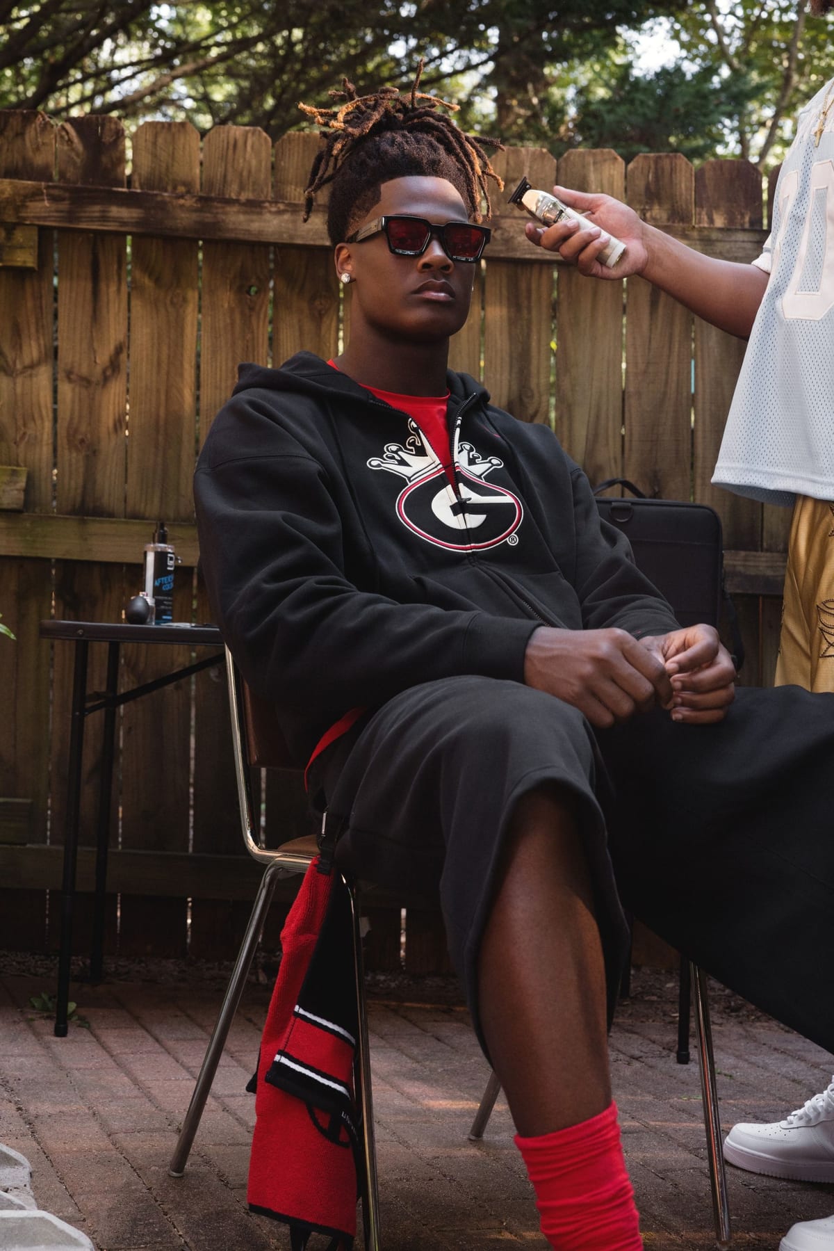 A Georgia player in dark sunglasses sits on a folding chair in a black hoodie printed with the Georgia G under a white crown, while someone works on his hair with clippers
