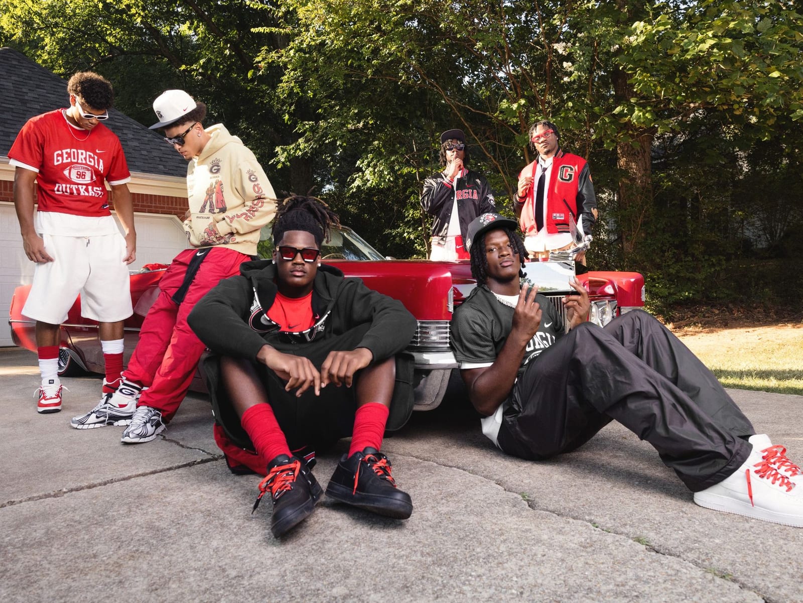 Six young men in the Georgia x OutKast collection in a driveway beside a red classic convertible: a red Georgia Outkast football jersey, a cream graphic hoodie, a red varsity jacket with black sleeves and a black satin bomber