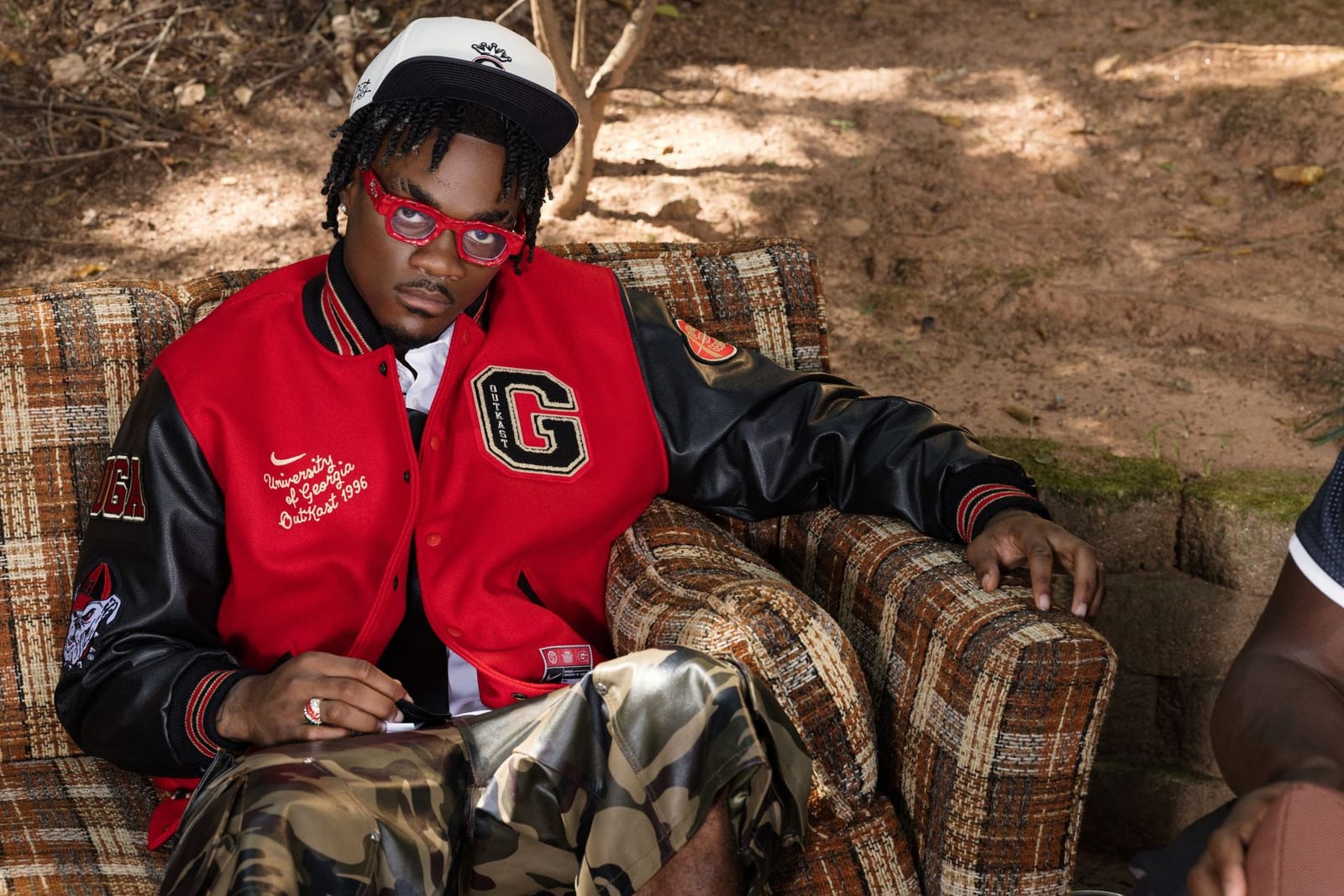 A man in red glasses and a white cap sits on a plaid couch in a red wool varsity jacket with black leather sleeves, a black chenille G on the chest and "University of Georgia Outkast 1996" stitched in white script
