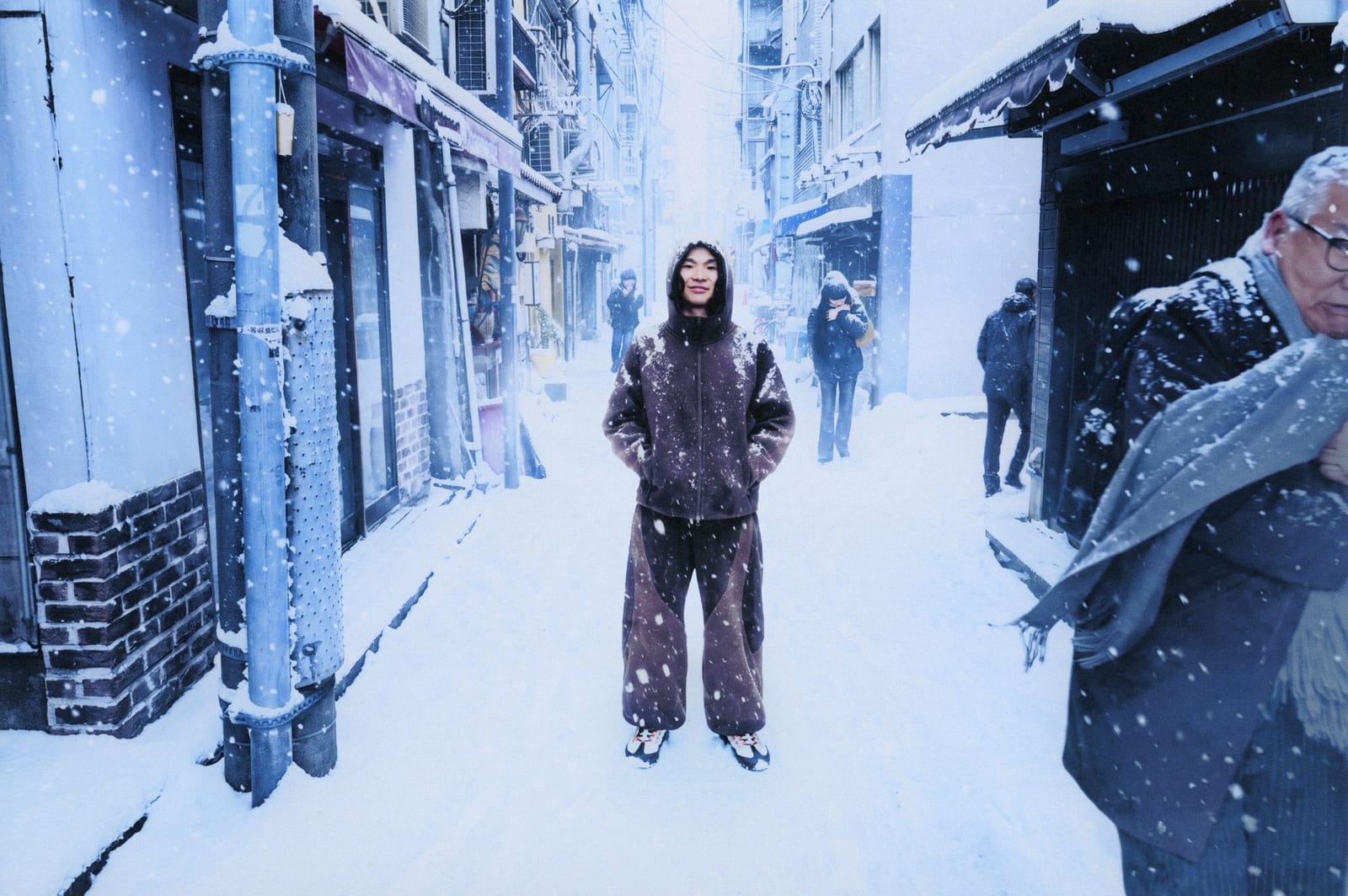 A man in a brown high-pile Nike Tech Boreas hooded top and matching trousers with darker panels stands in a snowy, narrow city street as snow falls, with an older man in a grey scarf at the right edge of the frame