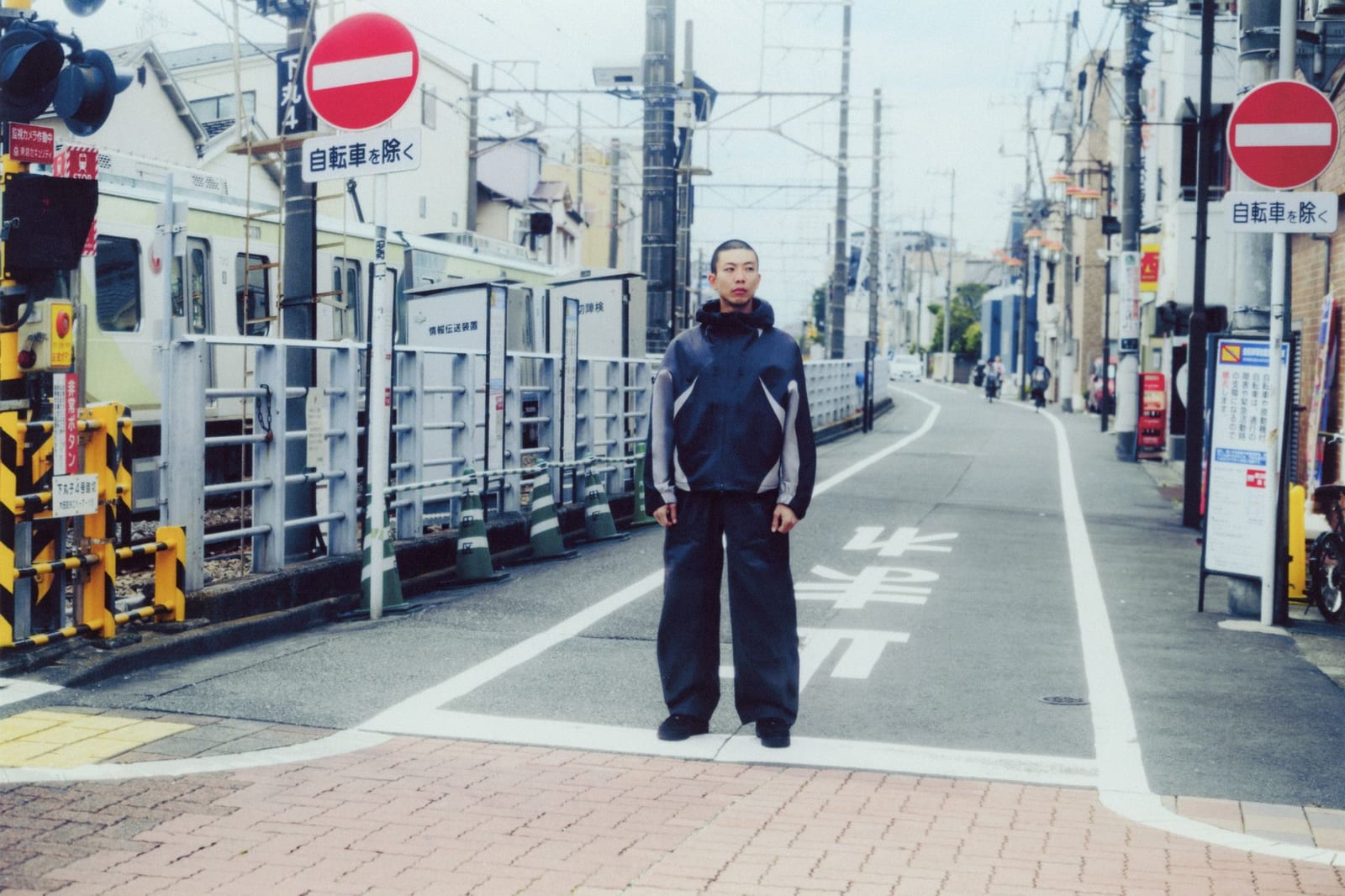 A man stands in the middle of a quiet Japanese street by a railway crossing in a black Nike Tech SF3 hooded jacket with curved grey side panels and dark wide-leg trousers