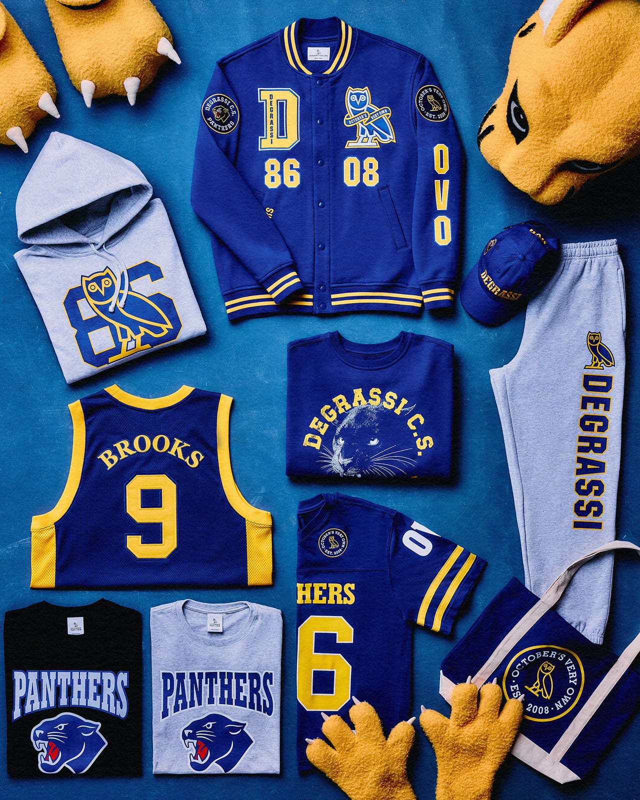 A flat lay of the OVO x Degrassi collection: a blue and yellow varsity jacket with OVO and Degrassi patches, a Brooks No. 9 basketball jersey, a Panthers No. 86 football jersey, an 86 hoodie, Panthers tees, a cap, sweatpants and a tote, next to a Panthers mascot costume