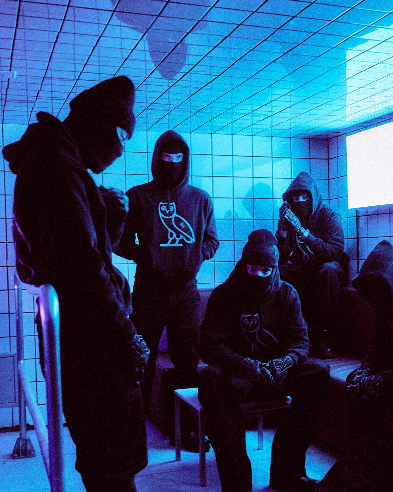 A group of people in black hoodies and balaclavas sitting and standing in a tiled room lit blue, one standing at the back with a large white OVO owl on the front of their hoodie