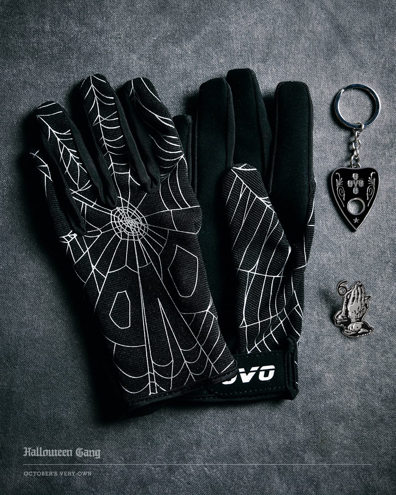 Black gloves printed with a white spider web and the OVO name, beside a silver and black planchette-shaped keychain and a silver praying-hands pin