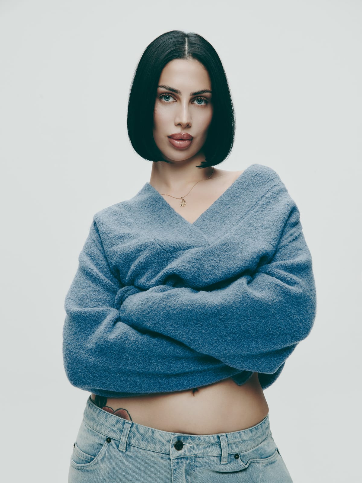 A woman with a black bob in a fuzzy dusty blue wrap sweater, arms crossed at her waist, with light blue jeans, against a pale grey wall