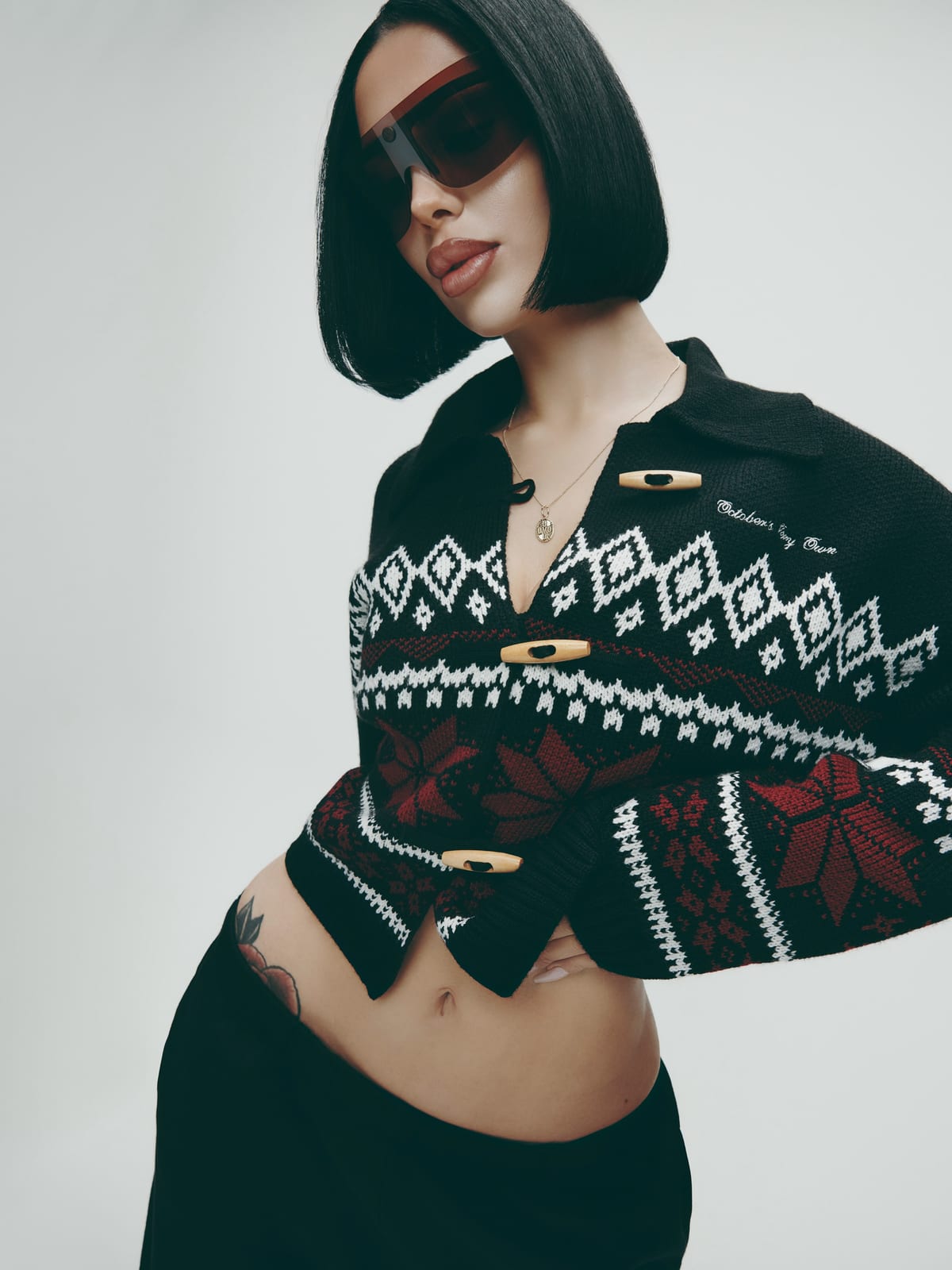 A woman with a black bob and red-tinted shield sunglasses in a cropped black Nordic-pattern cardigan with white diamonds, red snowflakes, toggle buttons and October's Very Own embroidered in white script, with a black skirt, against a pale grey wall