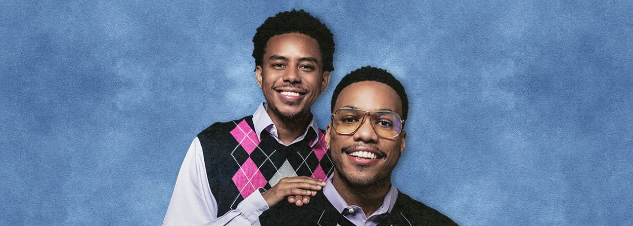 Anderson .Paak and Cordae in matching argyle vests against a mottled blue studio backdrop, both smiling at the camera