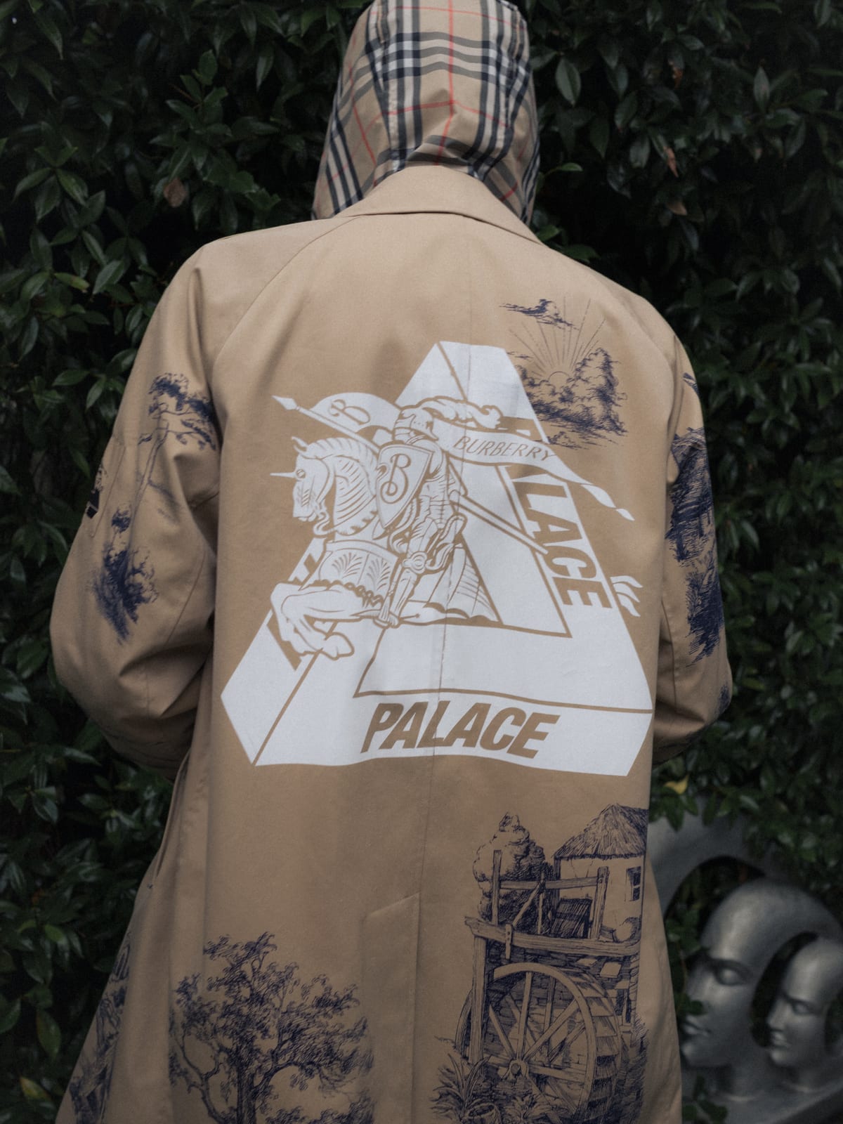 The back of a beige Palace x Burberry car coat, worn with a check hood up underneath, printed with a white Palace Tri-Ferg triangle holding Burberry's Equestrian Knight and navy drawings of trees, a cottage and a water wheel, against a hedge