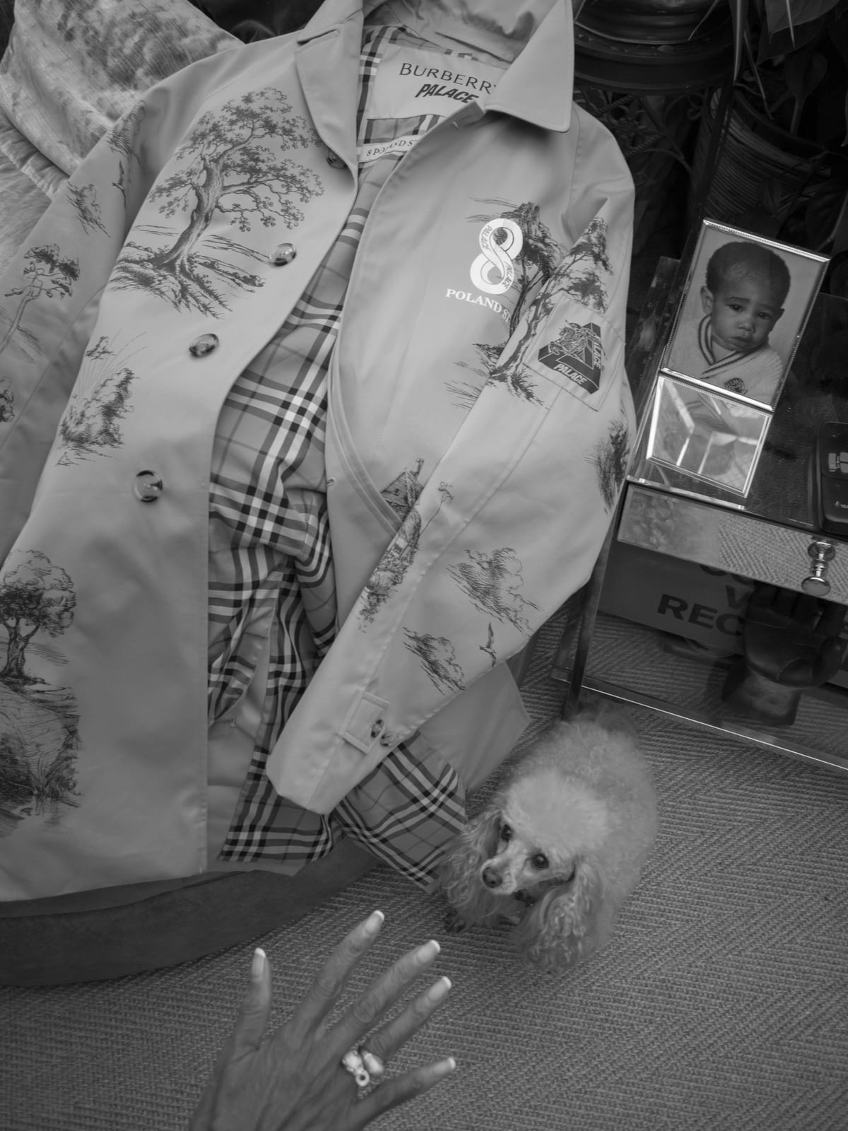 Black-and-white photo of the Palace x Burberry car coat laid open on a chair to show its check lining, its sleeves and body printed with trees, a label reading Burberry Palace and an 8 Poland St graphic on the chest, a poodle on the floor beside it