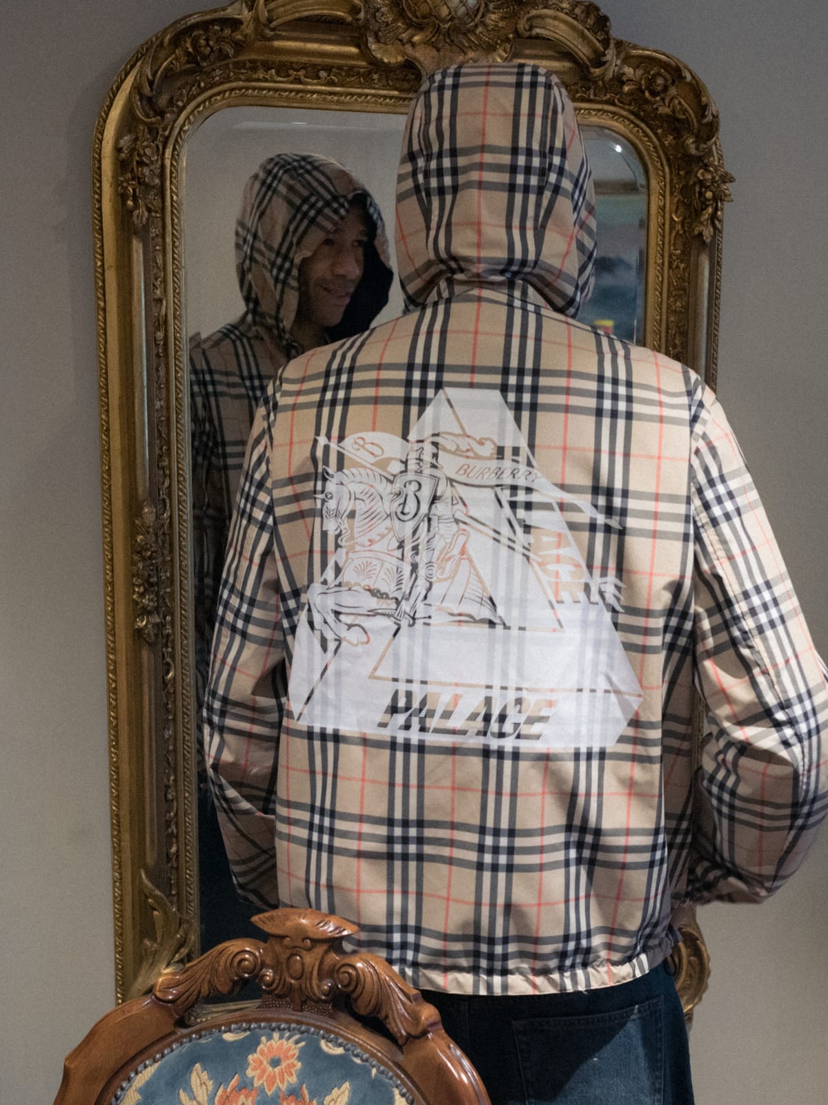 A man in a Palace x Burberry check hooded jacket with the hood up, seen from behind in front of a gilt mirror, a white Tri-Ferg and Equestrian Knight graphic across the back