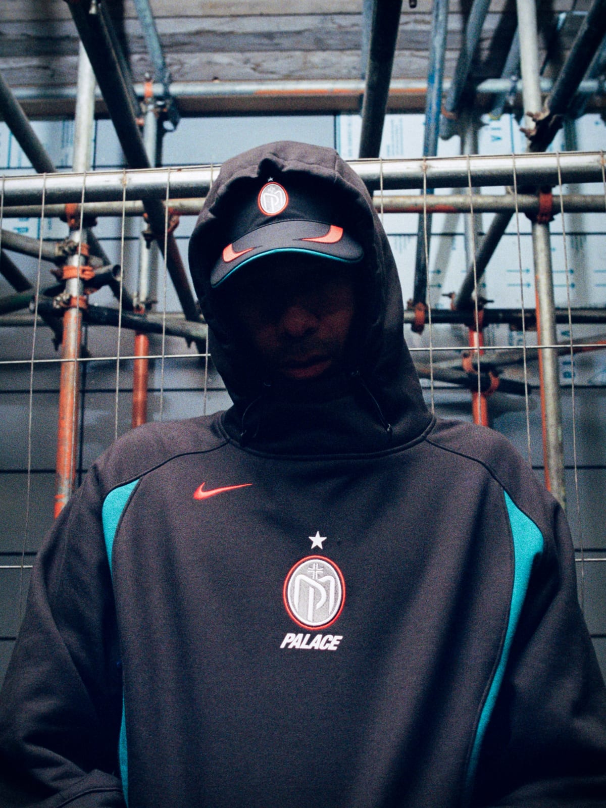 A man in a black Nike Swoosh cap with the hood of a black top pulled over it, the top with teal side panels, an orange Swoosh, a star over a round crest and PALACE on the chest, under scaffolding