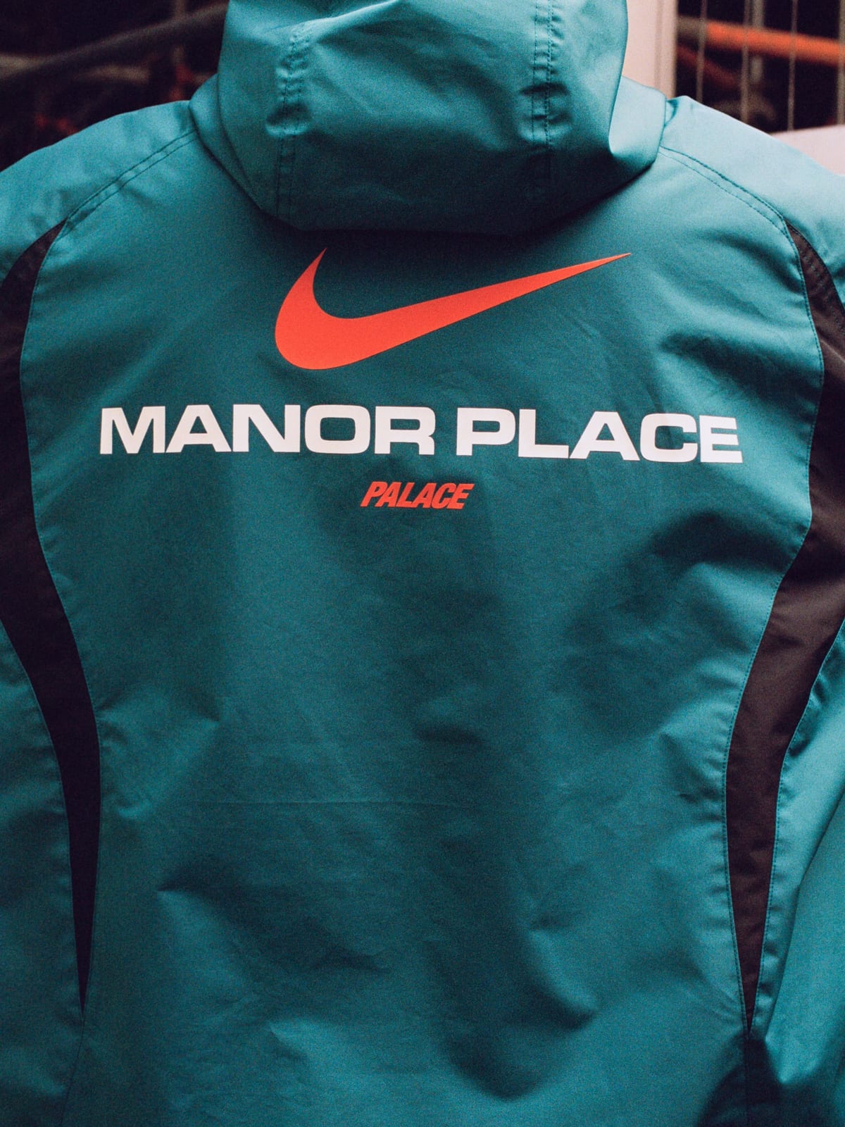 The back of a teal hooded zip jacket with dark side panels, printed with a large orange Nike Swoosh, MANOR PLACE in white capitals and Palace in orange