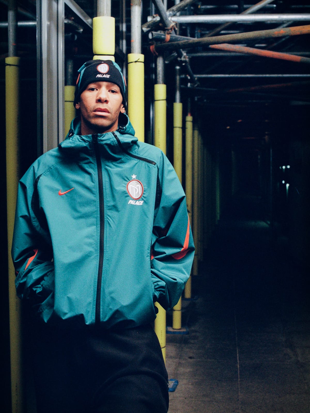A man in a black beanie stands under scaffolding in a teal hooded zip jacket with an orange Nike Swoosh, a round Manor Place crest and the Palace name on the chest