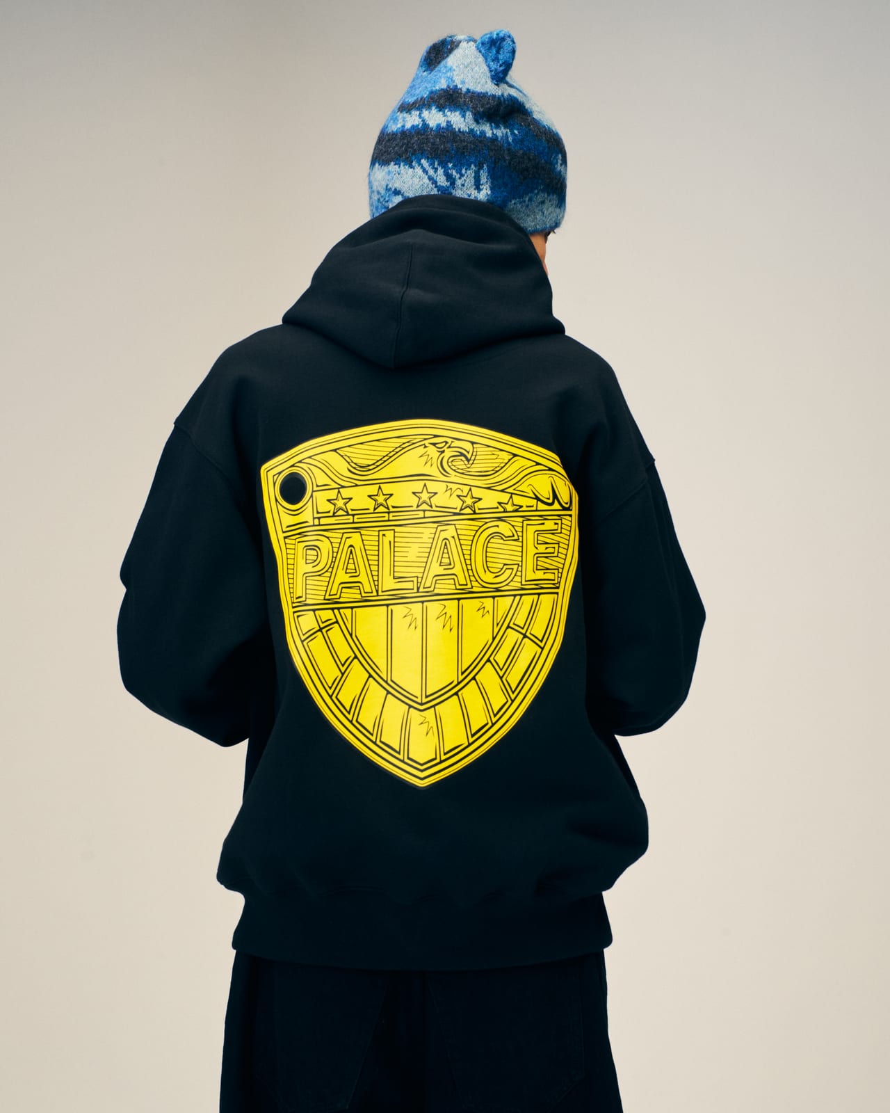 The back of a black Palace Winter 2026 hoodie with Judge Dredd's badge redrawn in yellow, with PALACE across it