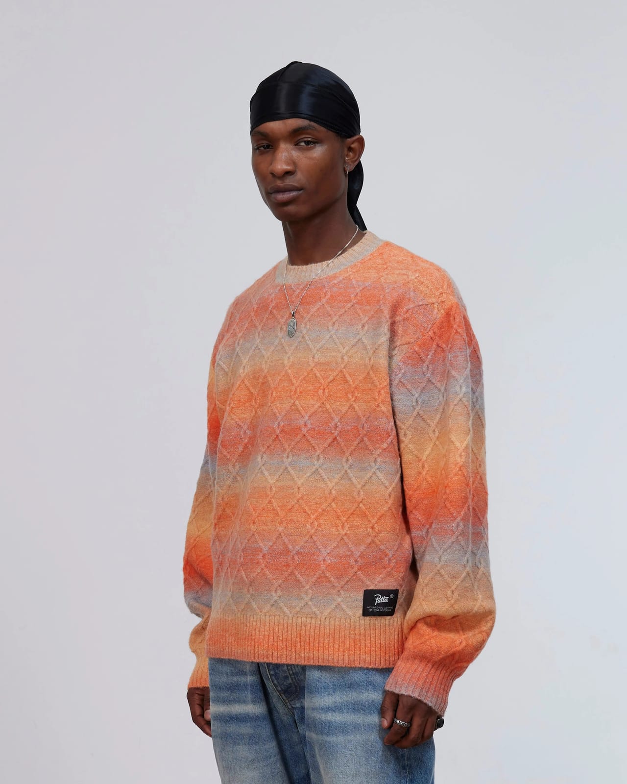 A model in a black durag wearing Patta's Space Dye Cable Knitted Sweater, an orange, peach and grey diamond cable knit with a black Patta label at the hem, over light blue jeans