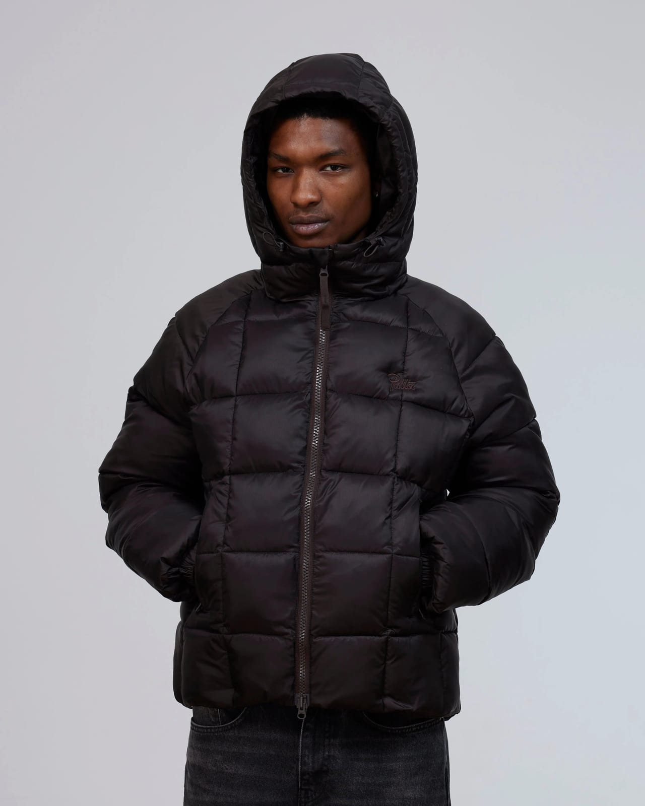 A model in Patta's hooded Square Quilted Puffer Jacket in dark brown, zipped up with the hood on and hands in the pockets, a small tonal Patta logo on the chest